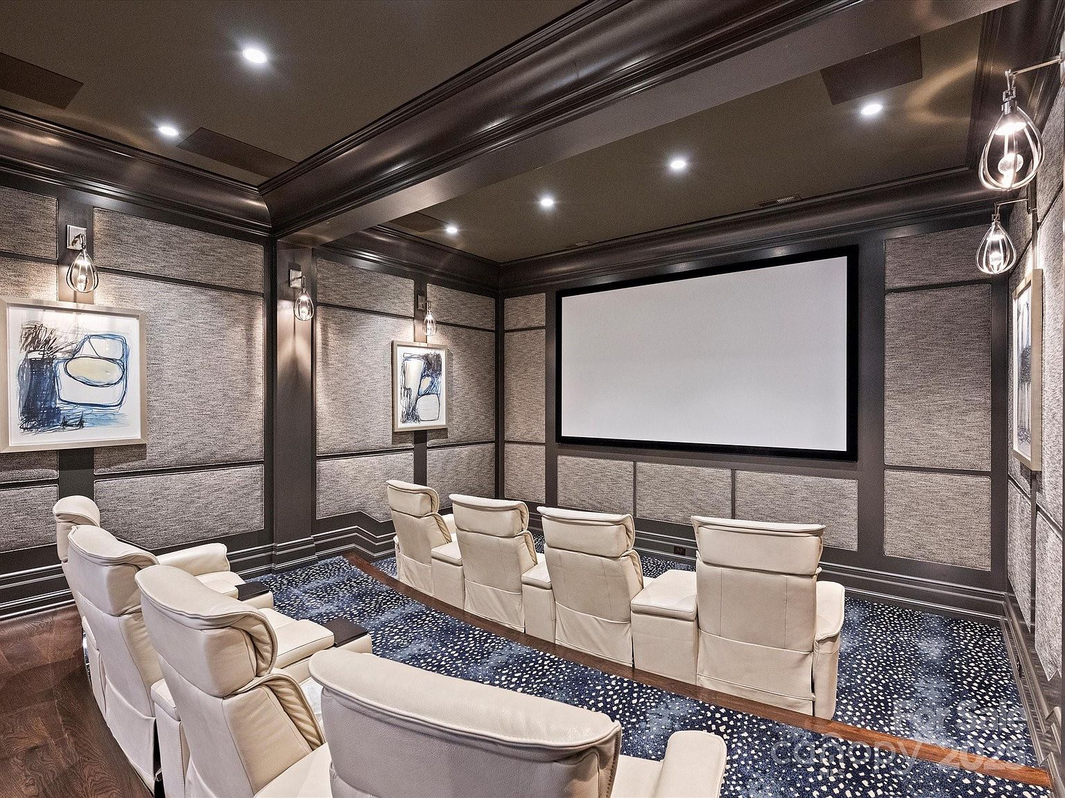 This dedicated home theater room features tiered rows of cream-colored leather reclining seats facing a large projection screen. The walls are adorned with fabric-paneled wainscoting and framed artwork, complemented by dark wood trim and recessed ceiling lighting. A patterned carpet adds texture to the space, creating a sophisticated and immersive cinematic environment.