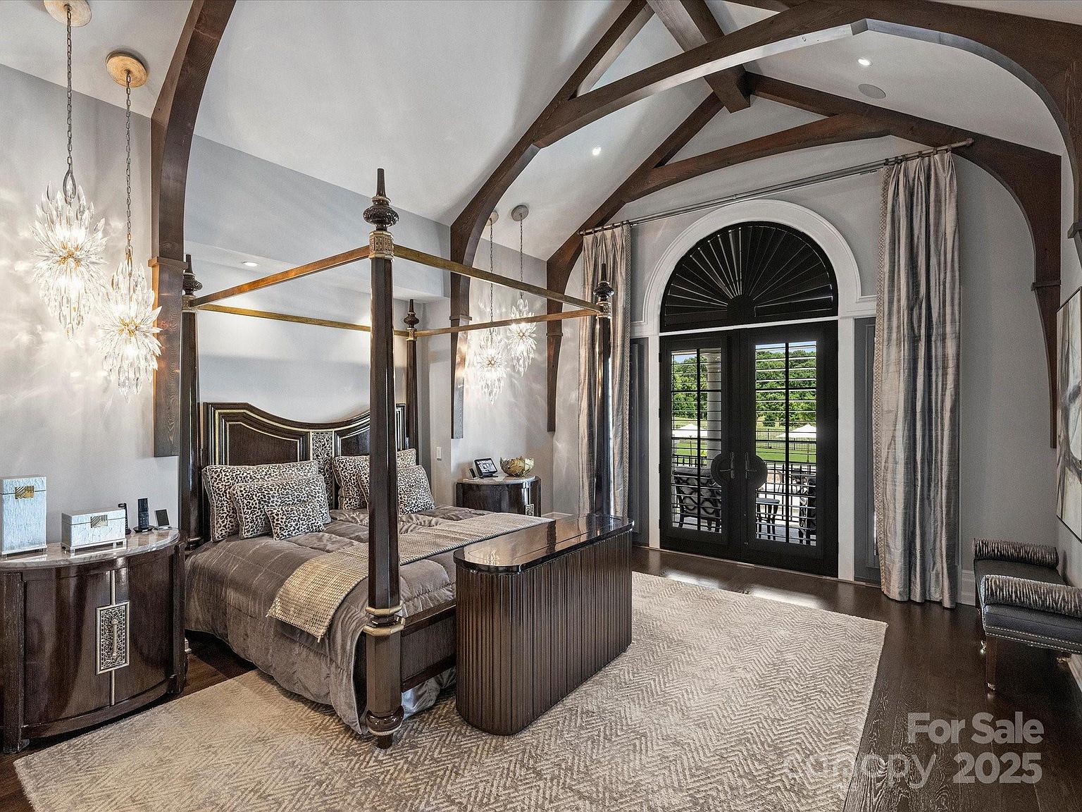 This luxurious primary bedroom features a dramatic vaulted ceiling with exposed dark wood beams and a grand four-poster bed as the centerpiece. A large arched glass door with black framing provides a view of the outdoors, flanked by elegant floor-length curtains. The room is accented by sophisticated crystal pendant lighting and a neutral patterned area rug, creating an atmosphere of refined elegance.