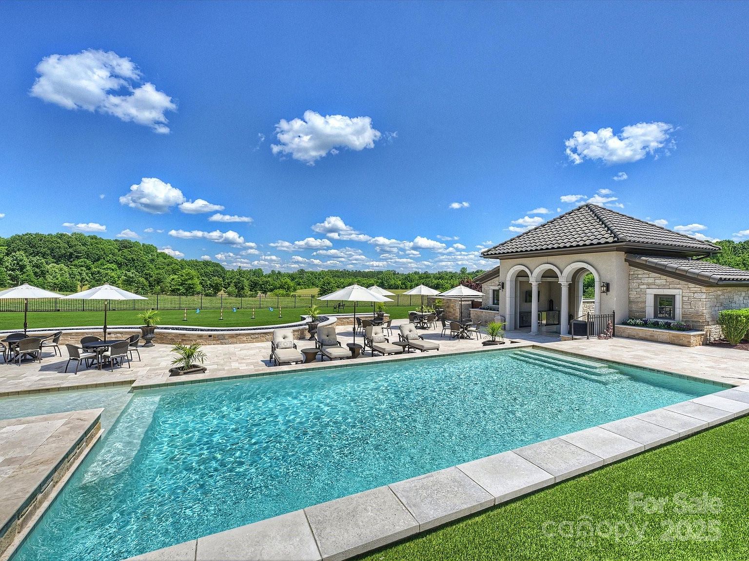 This expansive outdoor living area features a large, rectangular swimming pool with a shallow tanning ledge, set against a backdrop of lush green fields and a clear blue sky. A stone-clad pool house with arched entryways provides a shaded retreat, while the surrounding stone patio is furnished with multiple dining sets and lounge chairs under umbrellas. The scene captures a luxurious, resort-style backyard designed for relaxation and outdoor entertaining.
