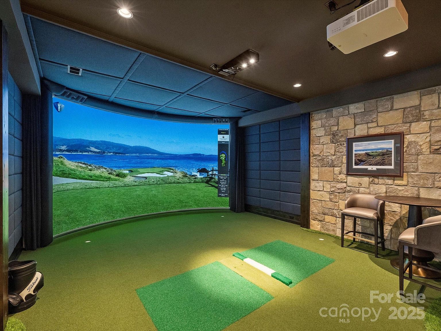 This dedicated home golf simulator room features a large, curved projection screen displaying a coastal golf course. The space is equipped with a professional-grade hitting mat, a ceiling-mounted projector, and a stone-clad wall accented by a framed golf course photograph. Two bar-style chairs and a small table are positioned to the side, creating a comfortable viewing area for guests.