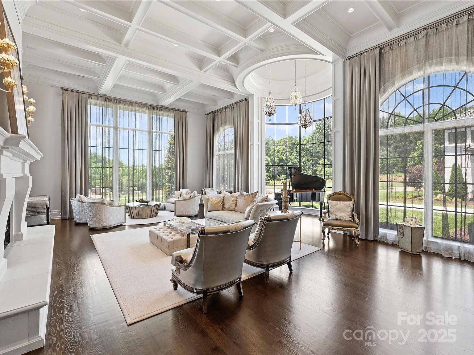 This expansive living room features a coffered ceiling and dark wood flooring, creating an elegant and formal atmosphere. The space is anchored by a large seating area with plush furniture and a central ottoman, while a grand piano sits near the tall, arched windows that offer views of the surrounding landscape. The room is bathed in natural light, highlighting the sophisticated decor and the architectural detail of the curved window alcove.