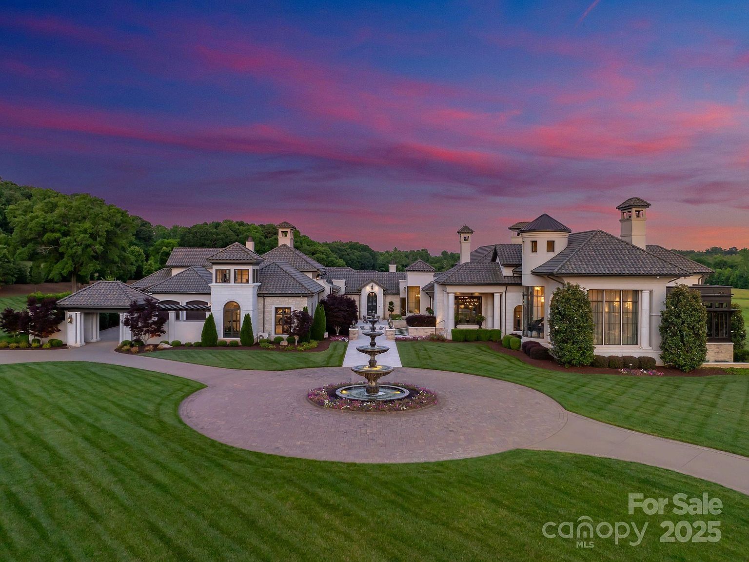 This aerial perspective captures a sprawling luxury estate at dusk, featuring a grand circular driveway centered around a multi-tiered stone fountain. The architecture showcases a sophisticated blend of light-toned masonry, dark-tiled roofs, and numerous chimneys, set against a vibrant, colorful sunset sky. Manicured green lawns and mature landscaping frame the residence, emphasizing its expansive scale and elegant curb appeal.