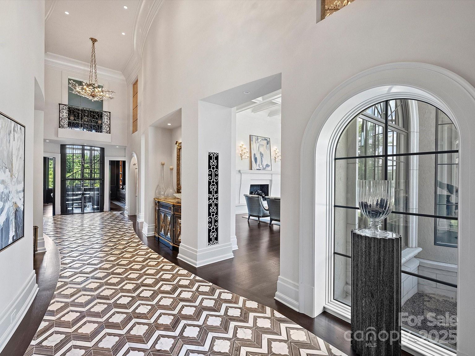 This grand foyer features a dramatic, high-ceilinged hallway with a striking patterned tile floor that leads the eye toward a distant living area. To the right, an arched window with black muntins provides natural light, while a decorative metal railing overlooks the space from an upper-level landing. The architectural design emphasizes scale and elegance, highlighted by a crystal chandelier and a sophisticated, neutral color palette.