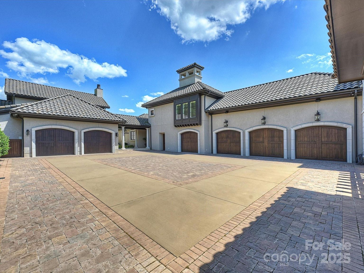 This expansive motor court features a multi-car garage configuration with five individual wooden carriage-style doors. The exterior showcases a sophisticated Mediterranean-inspired aesthetic with light-colored stucco walls, dark-framed windows, and a multi-gabled tile roof. The driveway is finished with a combination of smooth concrete and decorative brick pavers, creating a clean and upscale arrival area.