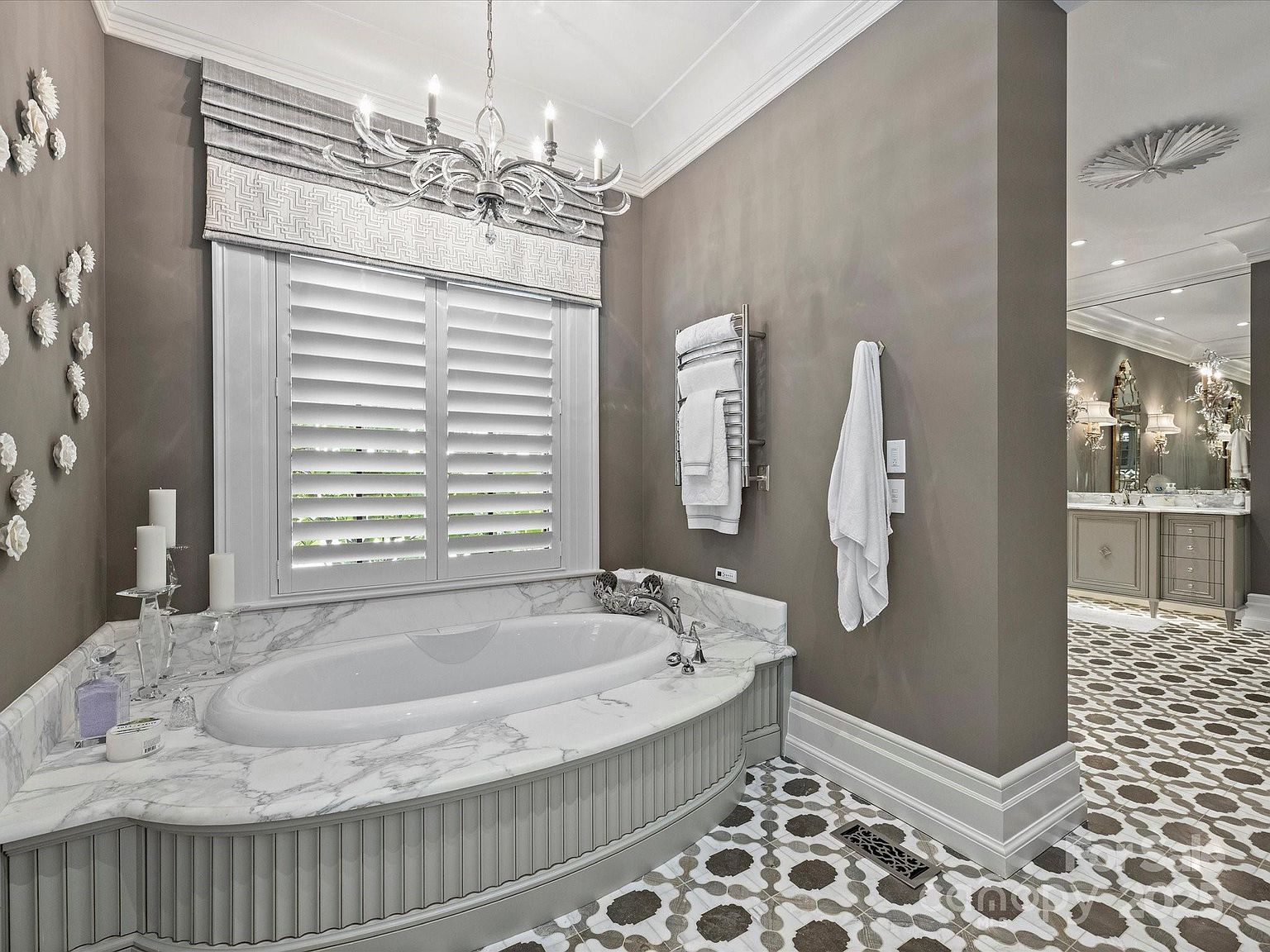 This elegant primary bathroom features a curved soaking tub set within a stone-topped vanity with decorative fluted paneling. The space is adorned with a crystal chandelier, wall-mounted floral accents, and a patterned tile floor that extends into the adjacent vanity area. A heated towel rack and shuttered windows complete the sophisticated, neutral-toned design.