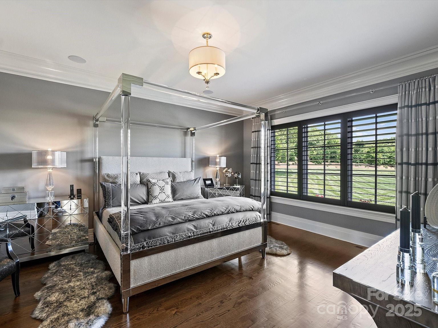 This elegant primary bedroom features a striking four-poster bed with a clear acrylic frame, centered against a soft gray wall. The room is appointed with mirrored nightstands, plush faux-fur rugs, and large windows with dark shutters that provide a view of the outdoor landscape. A sophisticated drum pendant light hangs from the ceiling, contributing to the room's polished and luxurious aesthetic.