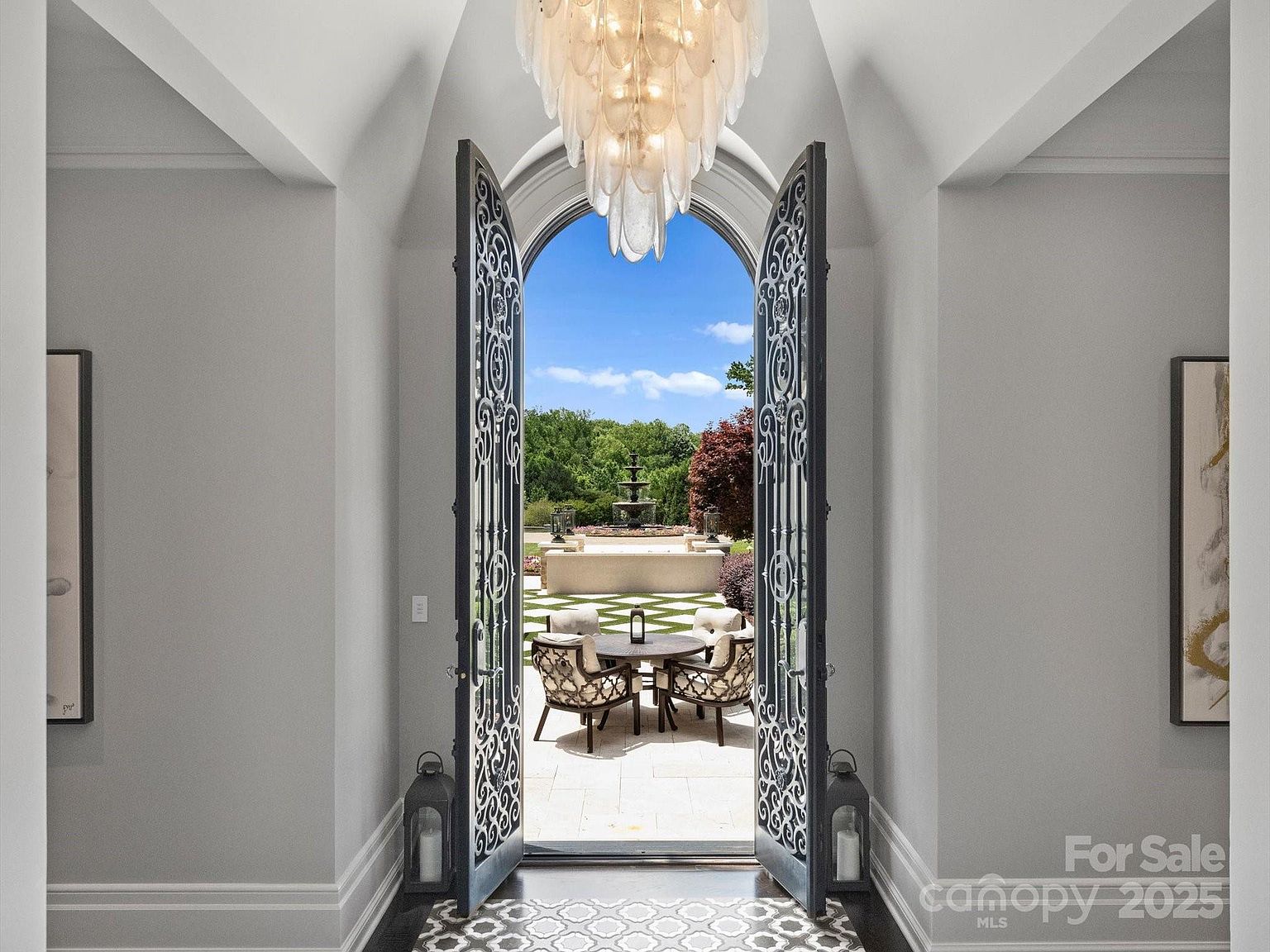This elegant entryway features a grand arched doorway with ornate metal-framed glass doors that open to a scenic outdoor patio and garden. A dramatic, multi-tiered crystal chandelier hangs from the vaulted ceiling, casting a sophisticated glow over the neutral-toned hallway. The perspective looks through the open doors toward a stone fountain and manicured landscaping, creating a seamless transition between the refined interior and the lush outdoor space.
