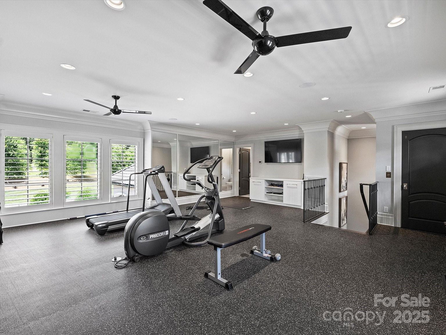 This spacious home gym features durable, speckled rubber flooring and is equipped with a treadmill, an elliptical machine, and a weight bench. The room is brightly lit by recessed lighting and two ceiling fans, with large windows along the left wall providing natural light and a view of the outdoors. A mirrored wall adds depth to the space, while a built-in white media console with a wall-mounted television offers entertainment options.