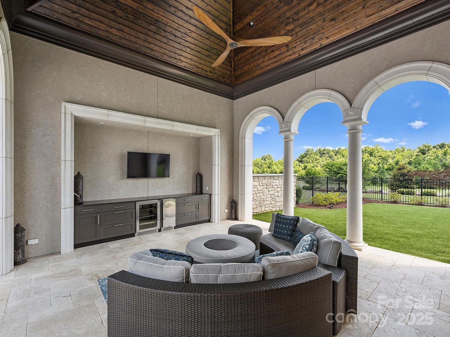 This luxurious covered outdoor living space features a high, wood-paneled vaulted ceiling with a large ceiling fan and elegant arched openings that frame views of the backyard. The area is furnished with a curved sectional sofa arranged around a central circular fire pit, complemented by a built-in cabinetry unit that includes a beverage cooler and television. The space is finished with light-toned stone flooring, creating a sophisticated and inviting atmosphere for outdoor entertaining.