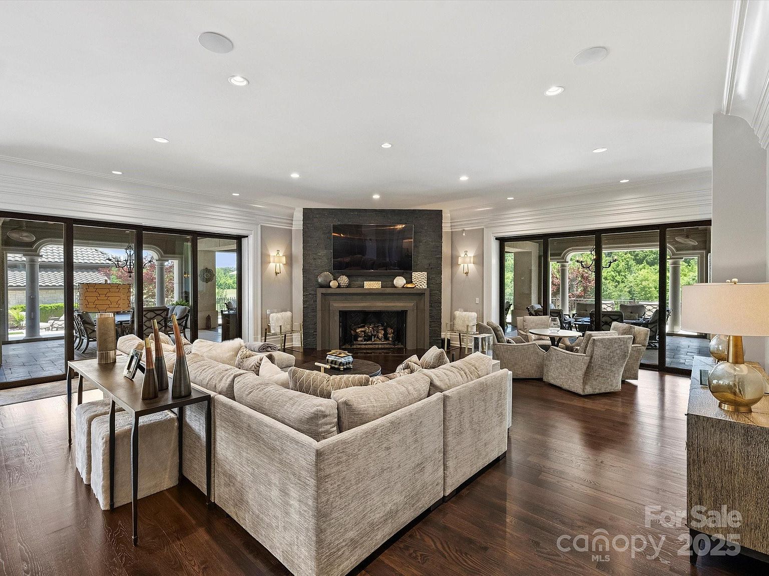 This spacious living room features a large, neutral-toned sectional sofa arranged around a central coffee table, facing a prominent fireplace with a dark stone-clad chimney breast. The room is framed by expansive glass sliding doors on both sides that provide views of the outdoor patio and lush greenery. Dark wood flooring runs throughout the space, complemented by recessed lighting and a sophisticated, open-concept layout.