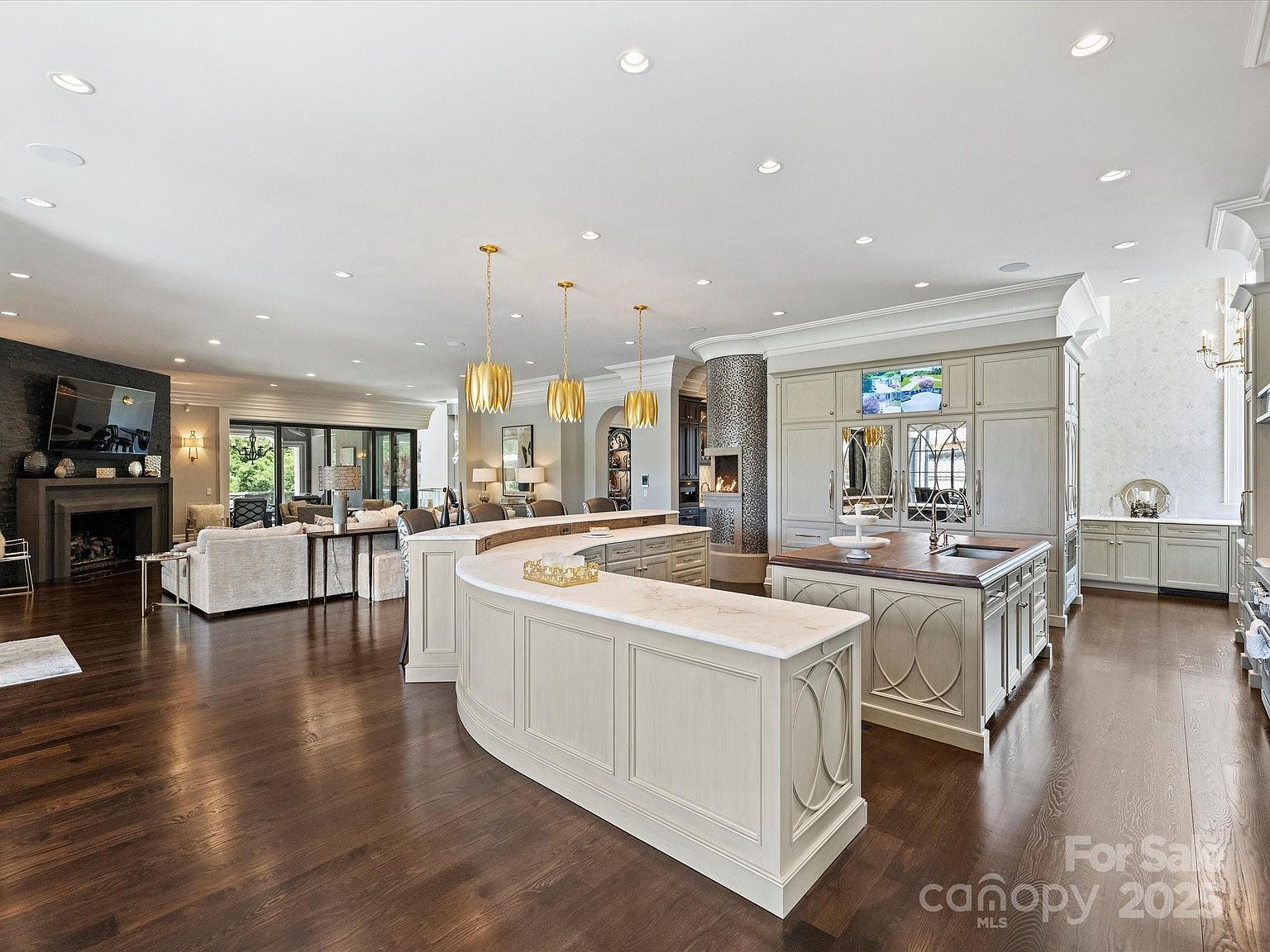 This expansive open-concept kitchen features a large, curved island with a light-colored stone countertop and three gold pendant lights overhead. Adjacent to the island is a secondary workspace with a dark wood countertop and integrated sink, set against a wall of light-toned cabinetry. The room flows seamlessly into a living area with a dark fireplace surround, creating a sophisticated and cohesive living space with dark hardwood flooring throughout.