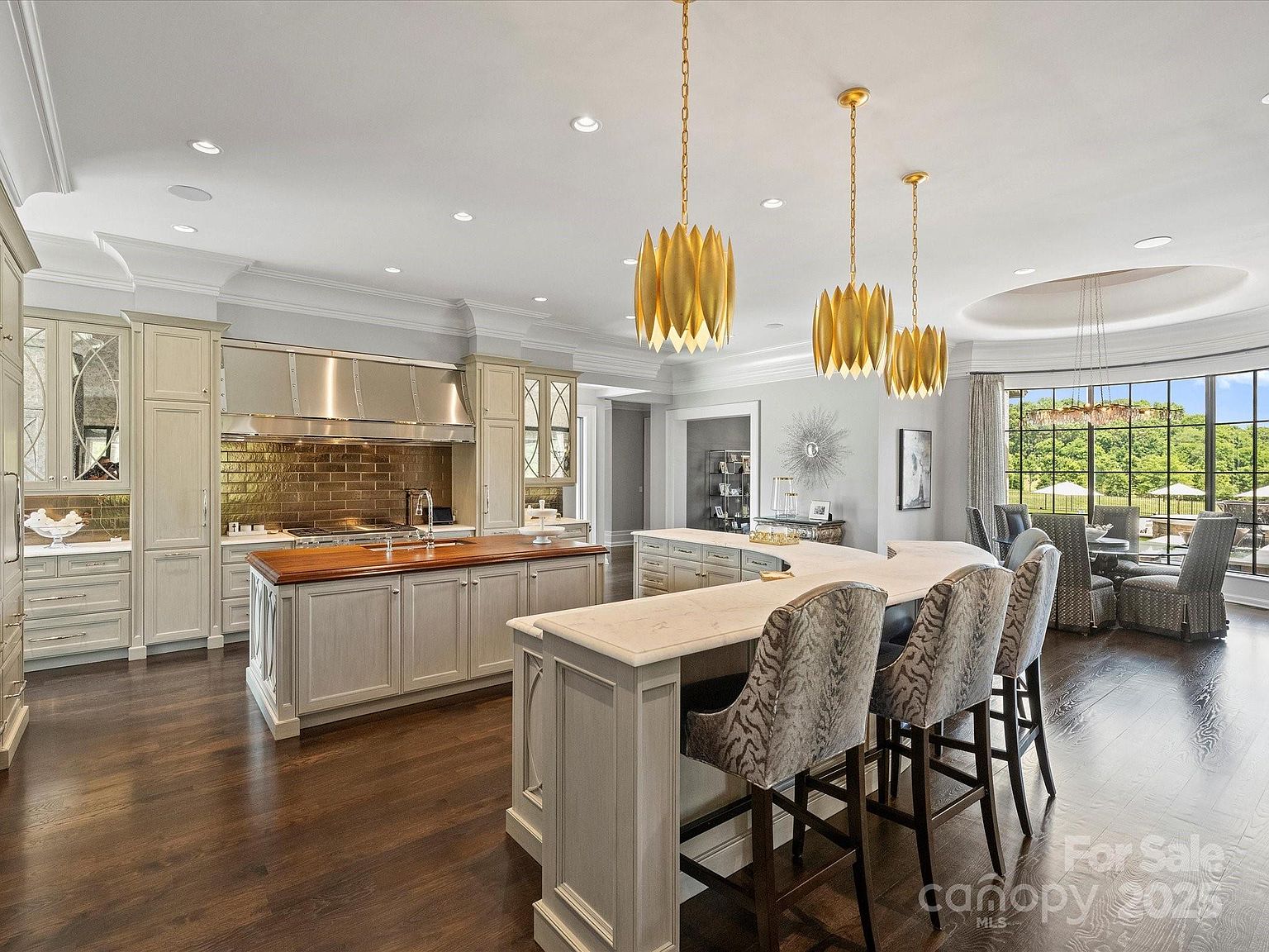 This expansive, high-end kitchen features a large central island with a warm-toned wood countertop and a secondary curved island with a light-colored stone surface. The space is illuminated by three distinctive gold-toned pendant lights and recessed lighting, while a large window wall provides a bright view of the outdoor landscape. The cabinetry is finished in a light, neutral tone, complemented by a metallic backsplash and professional-grade appliances, creating a sophisticated and airy atmosphere.
