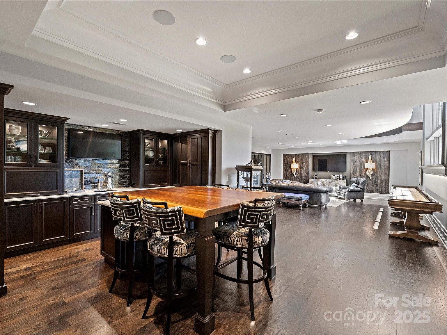 This expansive, open-concept basement features a large wet bar with dark cabinetry and a substantial wooden island with bar seating. The space transitions into a comfortable lounge area with a leather sofa and a game area equipped with a shuffleboard table. Recessed lighting and a tray ceiling design enhance the sophisticated, entertainment-focused atmosphere of this lower-level living space.