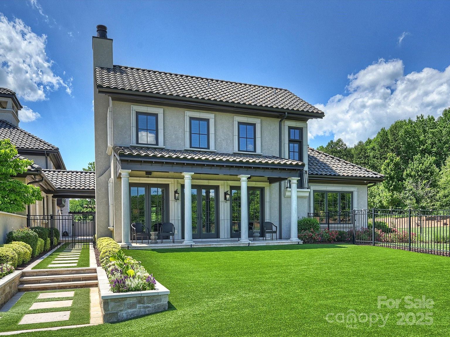 This rear view of a luxury residence showcases a sophisticated architectural style with a light-colored stucco facade, a dark tiled roof, and a covered patio supported by classic columns. The property features a manicured lawn, a stone-bordered garden bed with colorful flowers, and a paved walkway leading to a gated side entrance. The scene is framed by a black metal fence, creating a private and well-maintained outdoor living space.