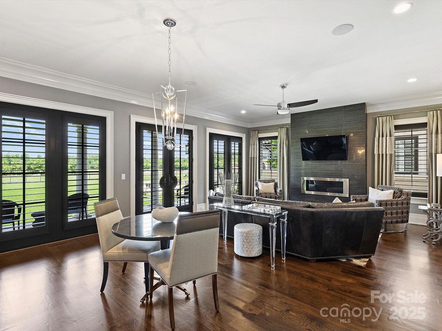 This open-concept living and dining area features dark hardwood flooring and a sophisticated, neutral color palette. A central seating area is anchored by a dark leather sofa facing a fireplace with a dark, textured surround and a mounted television. Large glass doors with black frames provide abundant natural light and views of the outdoor landscape, while a contemporary dining set sits in the foreground under a modern geometric chandelier.