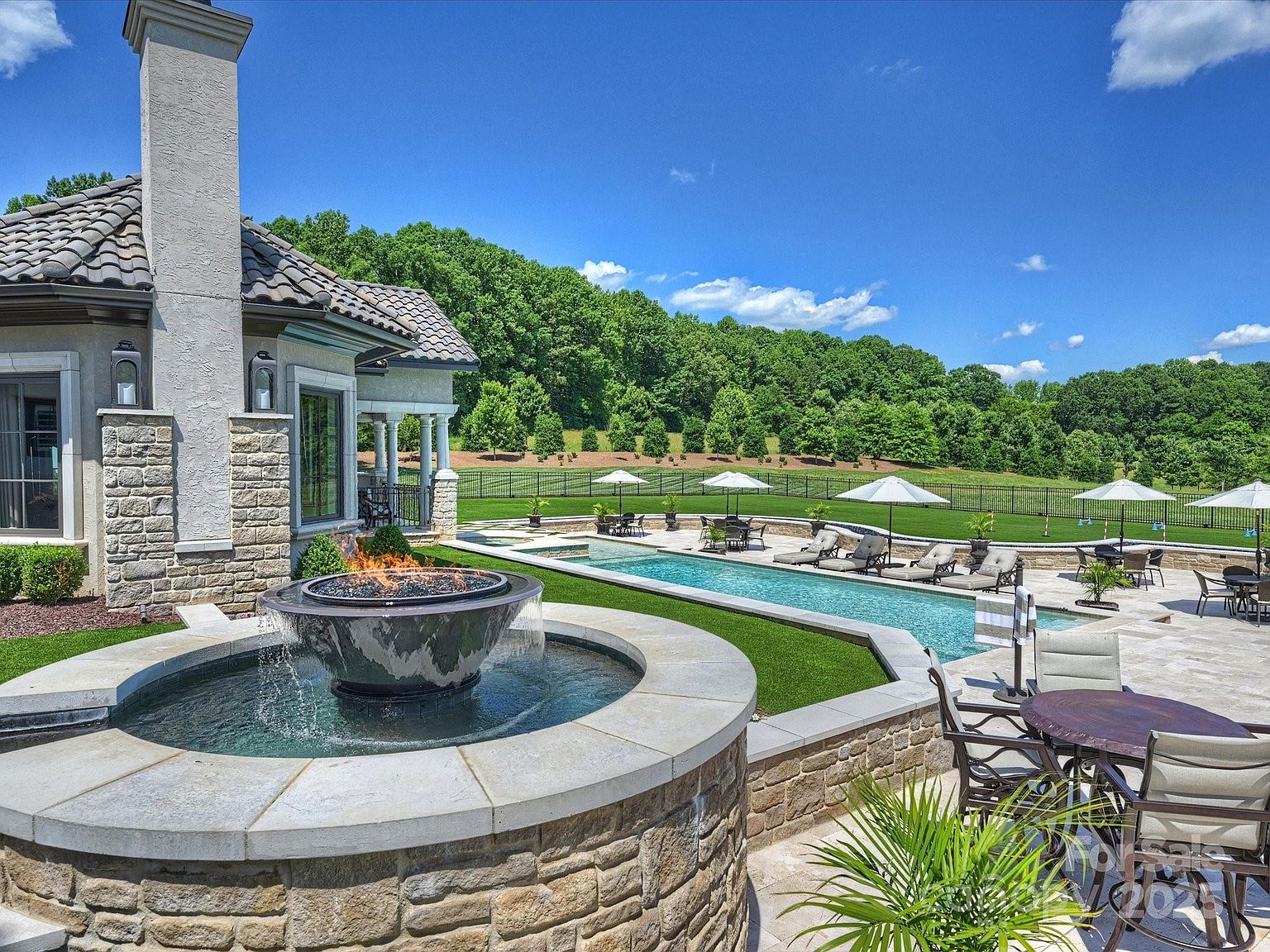 This expansive outdoor living space features a long, rectangular swimming pool bordered by a stone patio and a manicured lawn. In the foreground, a circular stone fire feature with a built-in fountain adds a sophisticated focal point, while multiple lounge chairs and dining sets are arranged under umbrellas for relaxation. The scene is framed by a stone-clad chimney breast on the house and a lush, tree-lined backdrop, creating a serene and luxurious atmosphere.
