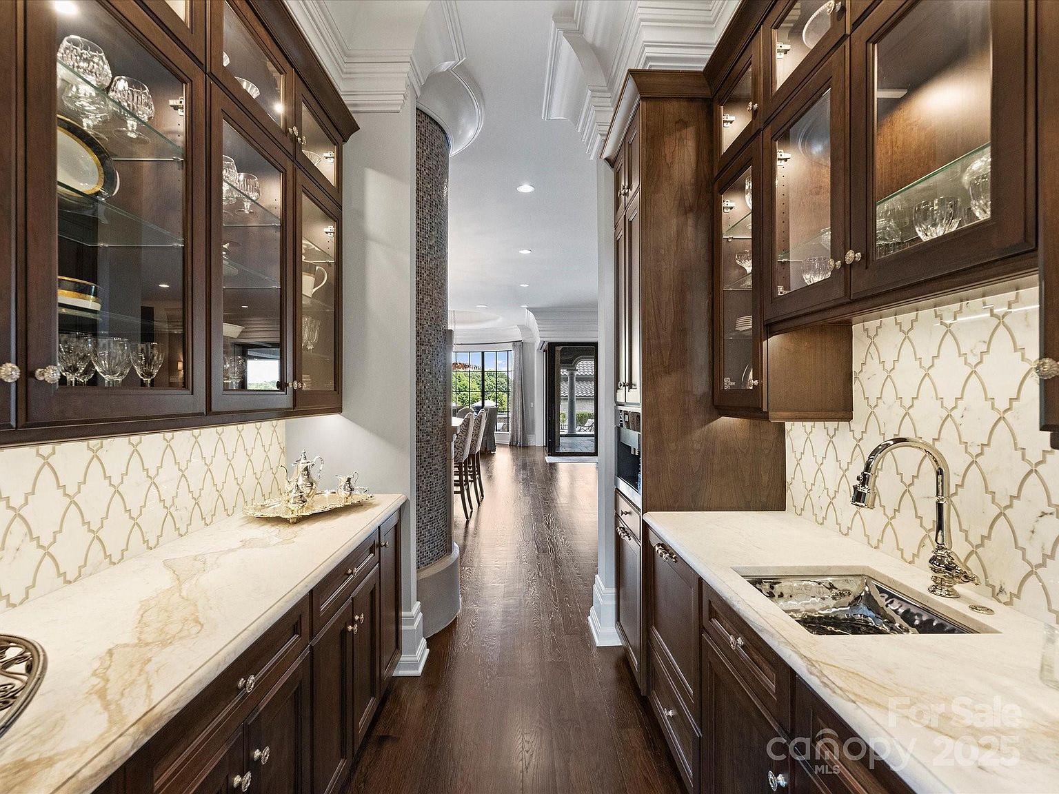 This elegant butler's pantry or wet bar features rich, dark wood cabinetry with glass-front upper cabinets and integrated lighting. The space is anchored by light-toned stone countertops, a decorative patterned tile backsplash, and a polished metal sink, creating a sophisticated transition area. The perspective looks through the galley-style layout toward a bright dining area, emphasizing the home's open flow and high-end finishes.