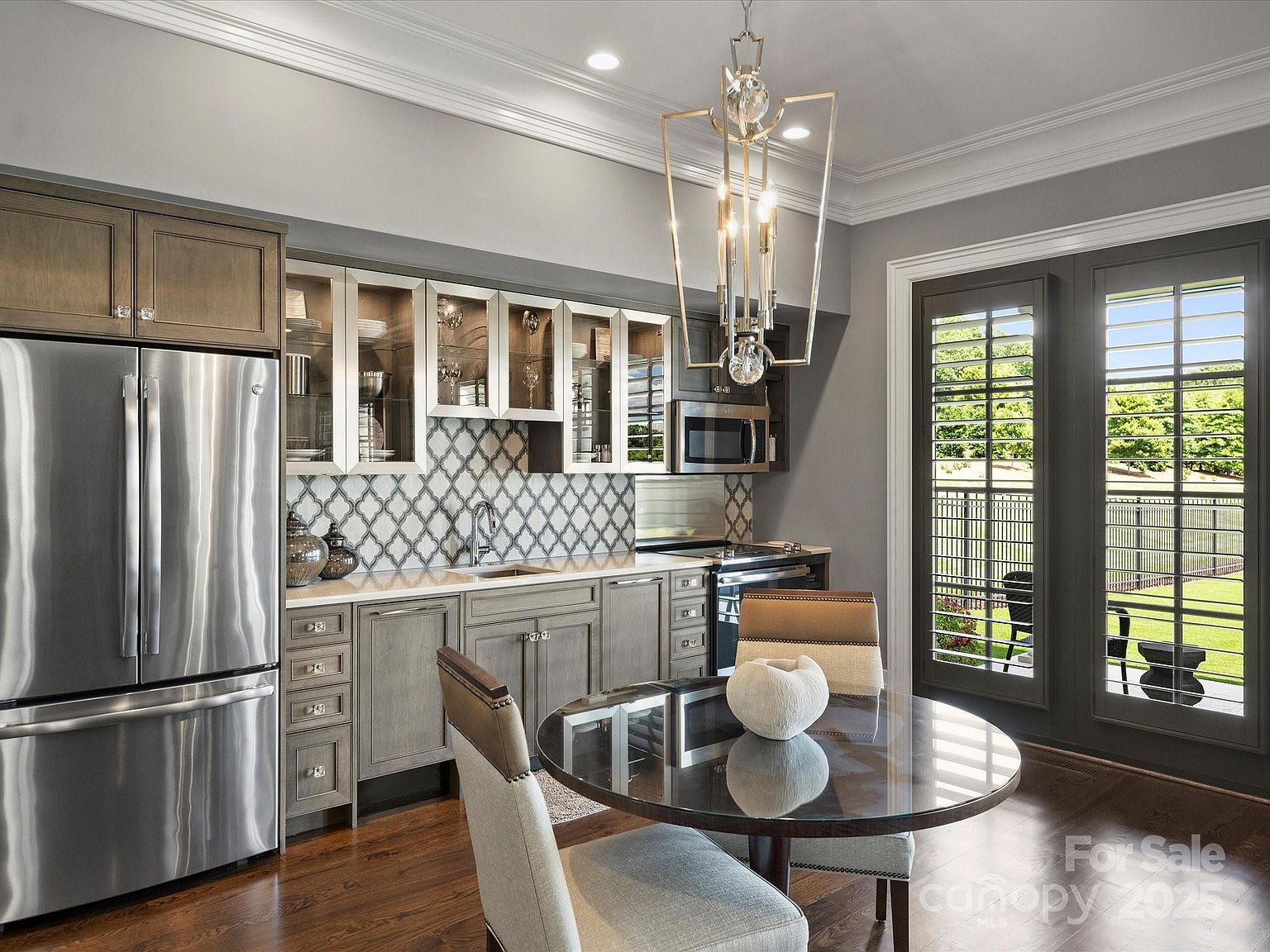 This elegant kitchen features a sophisticated blend of gray-toned cabinetry, stainless steel appliances, and a distinctive patterned tile backsplash. A round glass-top dining table with upholstered chairs sits in the foreground, while a modern geometric chandelier hangs above, creating a refined dining nook. The space is brightened by large glass doors with shutters that provide a view of the outdoor area, contributing to an airy and upscale atmosphere.