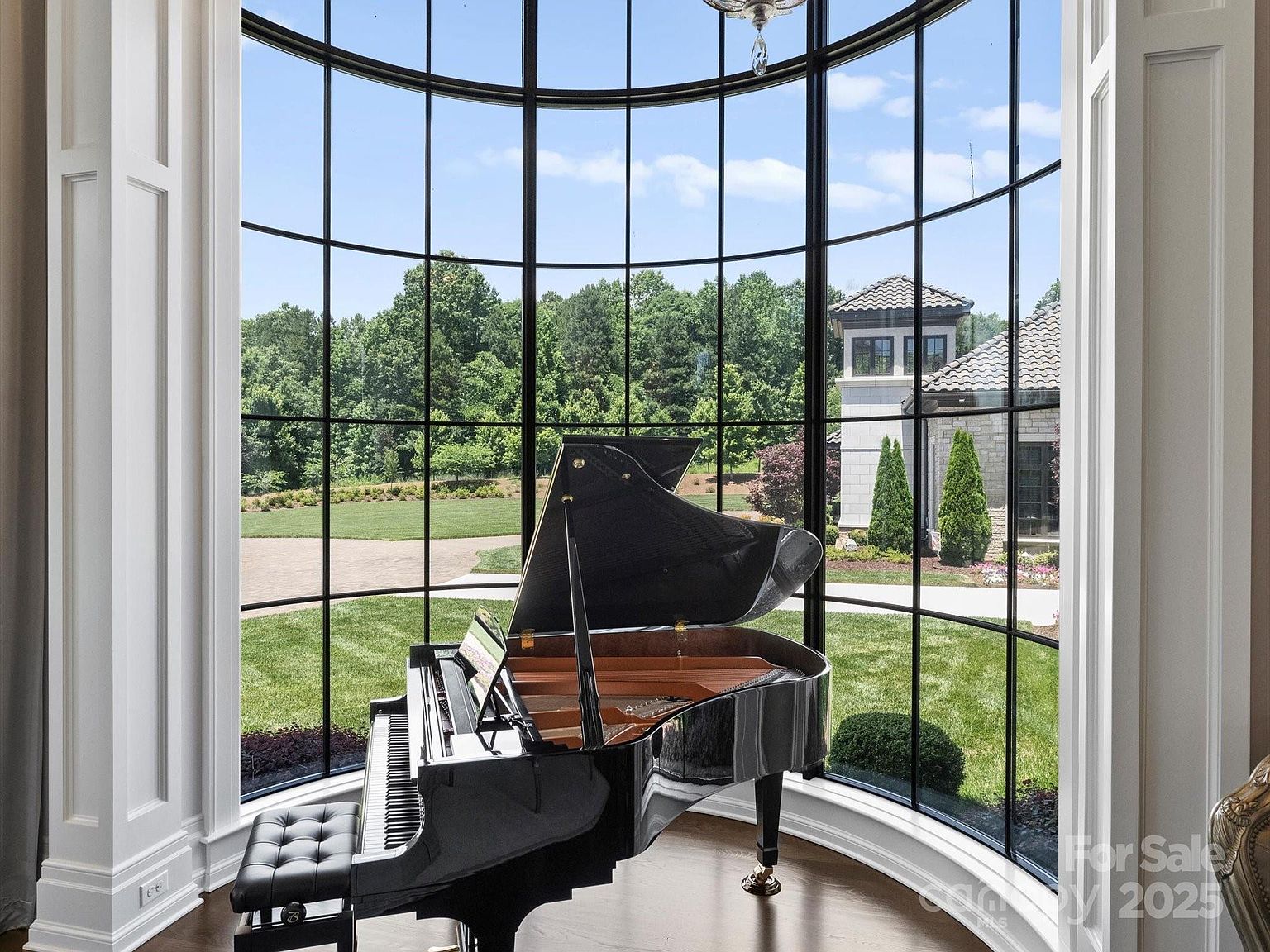 This elegant living space features a grand black piano positioned in front of a dramatic, curved wall of floor-to-ceiling windows that offer a panoramic view of the lush green lawn and neighboring estate. The room is framed by classic white architectural columns, creating a sophisticated and airy atmosphere. The perspective is centered on the piano, highlighting the seamless connection between the refined interior and the expansive outdoor landscape.