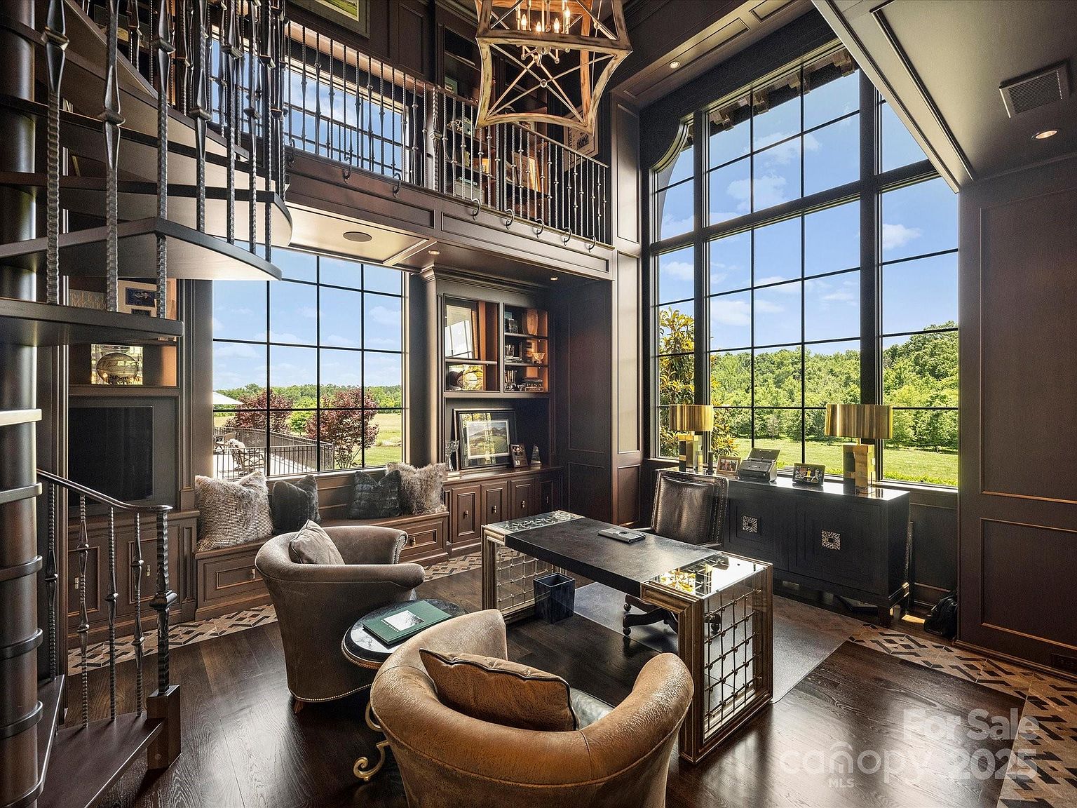 This sophisticated two-story home office features dark wood paneling, a spiral staircase, and a mezzanine level with a wrought-iron railing. The room is anchored by a modern desk with a metallic base and is illuminated by large, multi-pane windows that provide expansive views of the surrounding greenery. A built-in window seat with plush cushions and a geometric chandelier add to the luxurious, scholarly atmosphere of the space.
