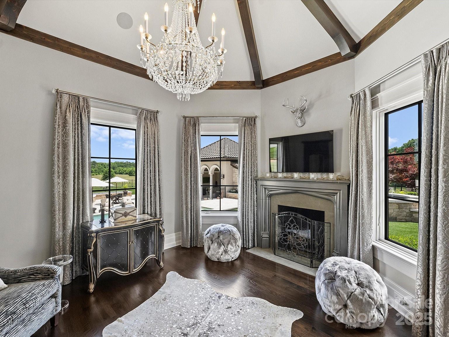 This elegant living room features a vaulted ceiling with exposed wood beams and a grand crystal chandelier as the central focal point. A fireplace with a decorative mantel serves as the anchor for the room, complemented by a mounted television and a silver deer head wall accent. The space is furnished with two plush, textured ottomans, a patterned area rug, and a dark wood cabinet, all set against a backdrop of tall windows draped in soft, patterned curtains.