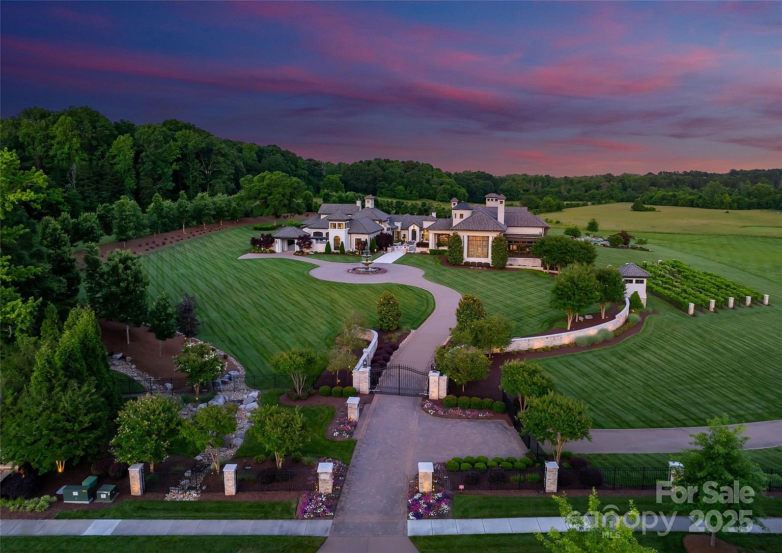 This expansive aerial view captures a grand estate set against a dramatic sunset sky. The property features a long, winding paved driveway leading through a gated entrance to a large circular motor court and a sprawling, multi-gabled residence. The meticulously manicured grounds include vast green lawns, mature trees, and a structured garden area, all framed by a peaceful rural landscape.