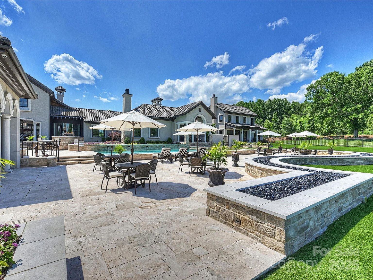 This expansive stone-paved patio serves as a luxurious outdoor living space, featuring a large swimming pool and multiple seating areas shaded by umbrellas. A prominent, curved stone-walled fire feature filled with dark glass rocks anchors the foreground, while the rear of the grand residence showcases traditional architectural details and multiple chimneys. The scene is captured from a low-angle perspective, emphasizing the vastness of the manicured lawn and the seamless transition between the home's interior and the outdoor entertainment area.