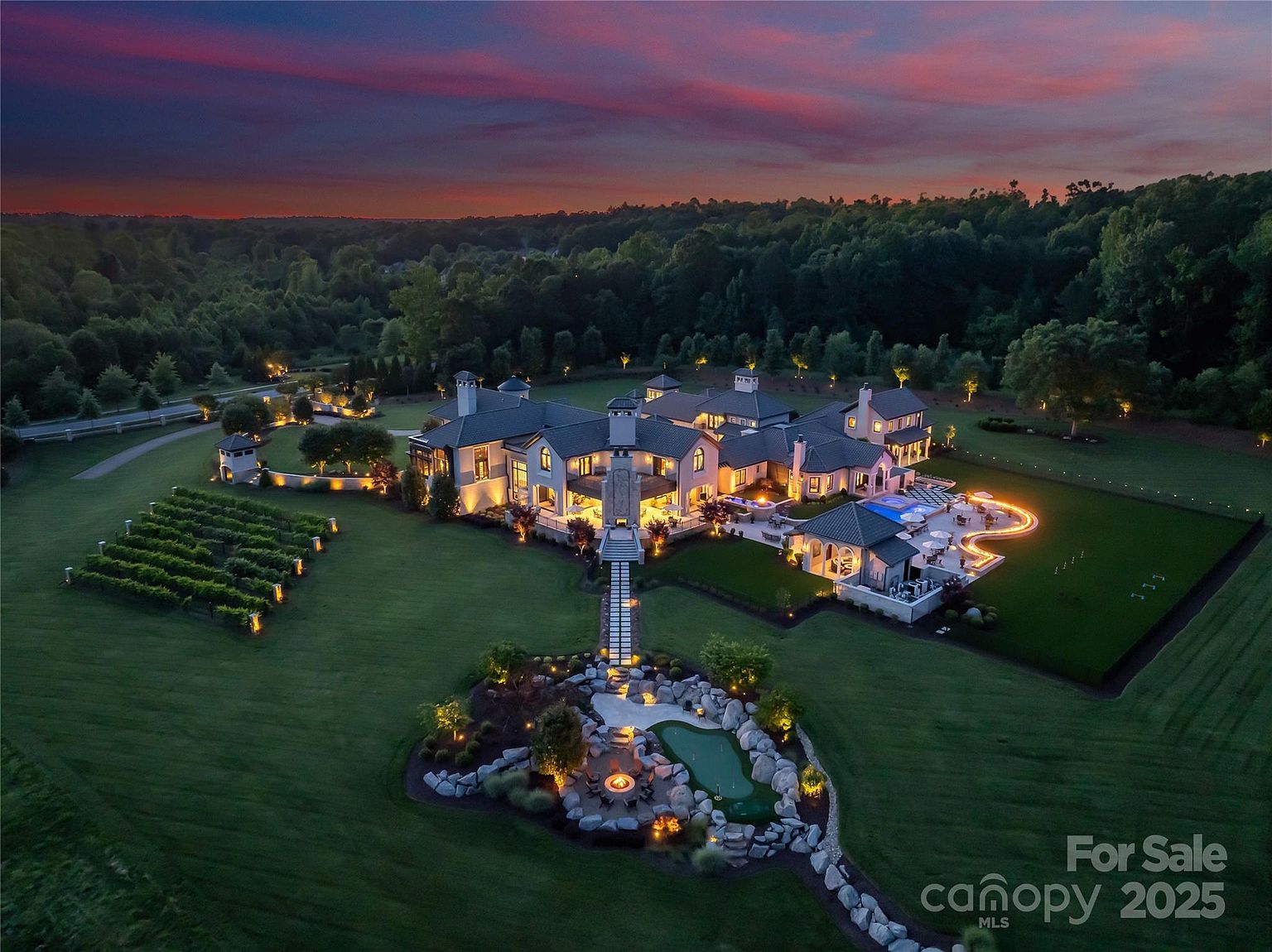 This stunning aerial view captures a sprawling luxury estate at dusk, set against a vibrant sunset sky. The property features an expansive main residence with multiple rooflines, a private vineyard, a stone-accented fire pit area with a small putting green, and a resort-style pool deck. The meticulously landscaped grounds are illuminated by warm exterior lighting, creating an inviting and sophisticated atmosphere.