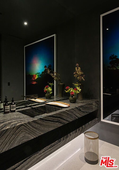 This sophisticated, moody bathroom features a striking, dark-veined marble vanity that serves as the centerpiece of the room. The space is accented by vibrant floral arrangements and large, dramatic art pieces that reflect in the mirror, creating a luxurious and artistic atmosphere. The minimalist design is enhanced by soft, under-cabinet lighting and a sleek, modern wastebasket, emphasizing a high-end, contemporary aesthetic.