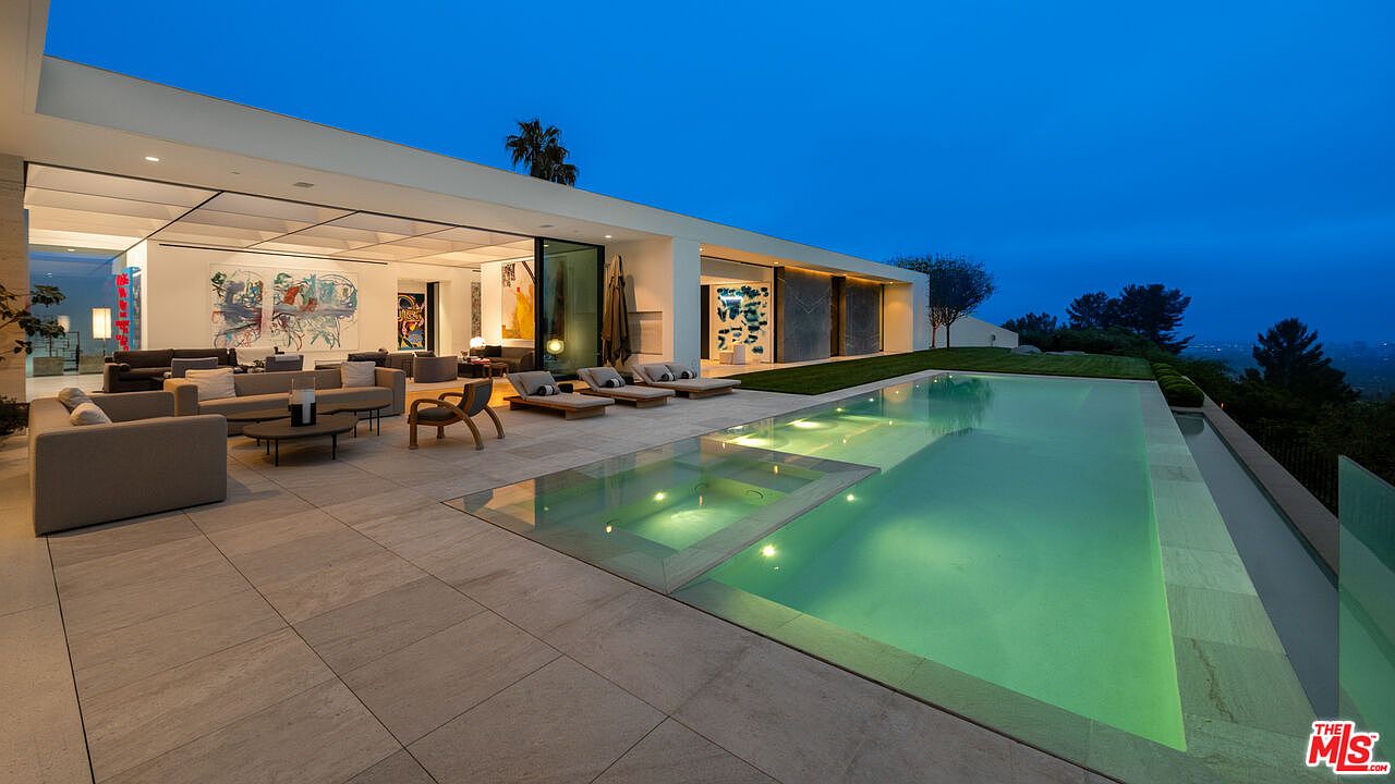 This stunning modern luxury estate features a seamless indoor-outdoor living space that opens onto a sprawling stone patio with a sleek, illuminated infinity-edge pool and integrated spa. The architecture emphasizes clean lines and expansive glass walls, creating a sophisticated atmosphere perfect for evening entertaining. The perspective captures the serene transition from the warm, art-filled interior living area to the cool, blue-toned twilight landscape overlooking the city lights.