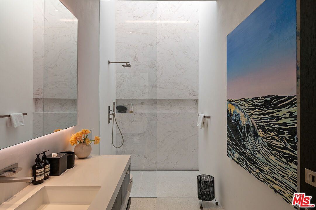 This modern, minimalist bathroom features a sleek vanity with a stone countertop, a large wall-to-wall mirror, and a spacious walk-in shower with marble-tiled walls. A striking piece of wave-themed artwork hangs on the wall, adding a vibrant pop of color to the neutral, sophisticated space. The room is bathed in natural light, creating a serene and high-end spa-like atmosphere.