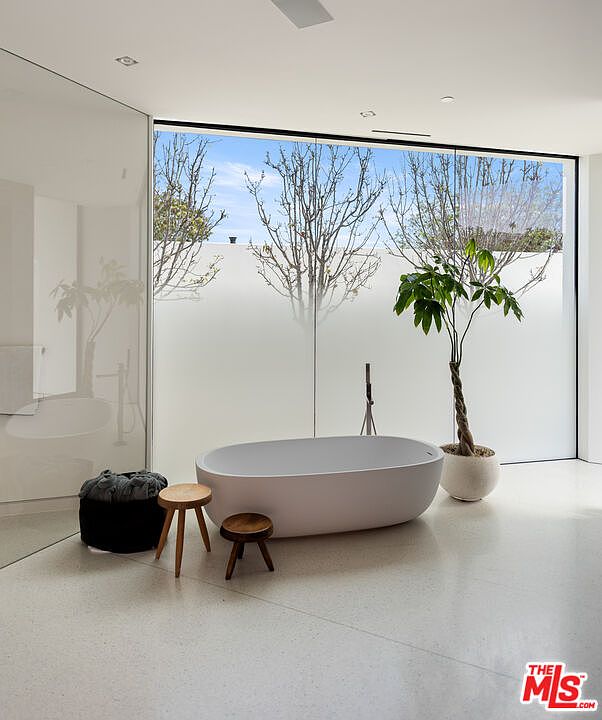 This minimalist primary bathroom features a sleek, freestanding oval soaking tub positioned in front of a large, floor-to-ceiling frosted glass window that allows for soft, diffused natural light. The space is accented by a potted money tree, two small wooden stools, and a black storage basket, all set against a clean, light-colored terrazzo floor. The overall aesthetic is serene, modern, and spa-like, emphasizing a tranquil atmosphere.