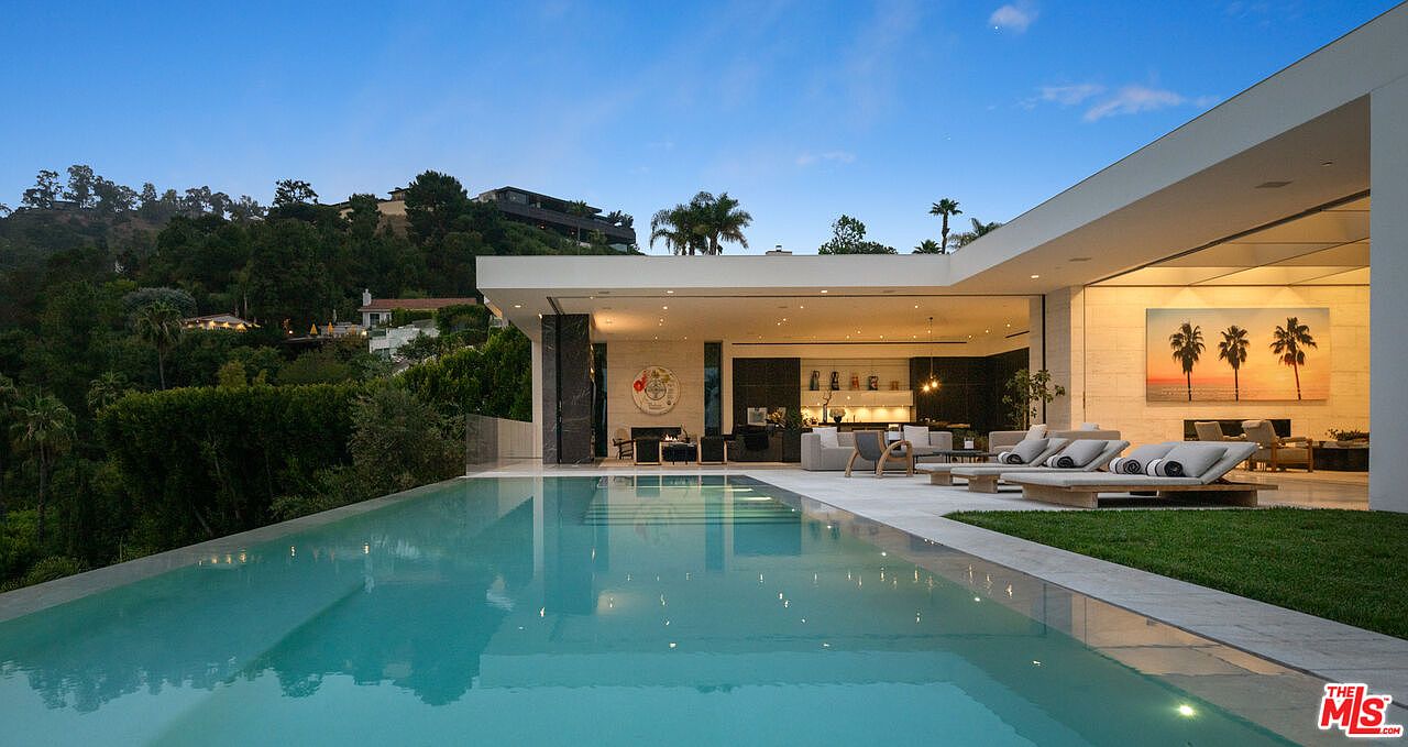 This stunning modern luxury estate features a seamless indoor-outdoor transition, highlighted by a large infinity-edge pool that reflects the clear blue sky. The expansive patio area is furnished with sleek lounge chairs and overlooks a lush, hilly landscape, creating a serene and sophisticated atmosphere. The architecture emphasizes clean lines and open-concept living, perfect for high-end entertaining.