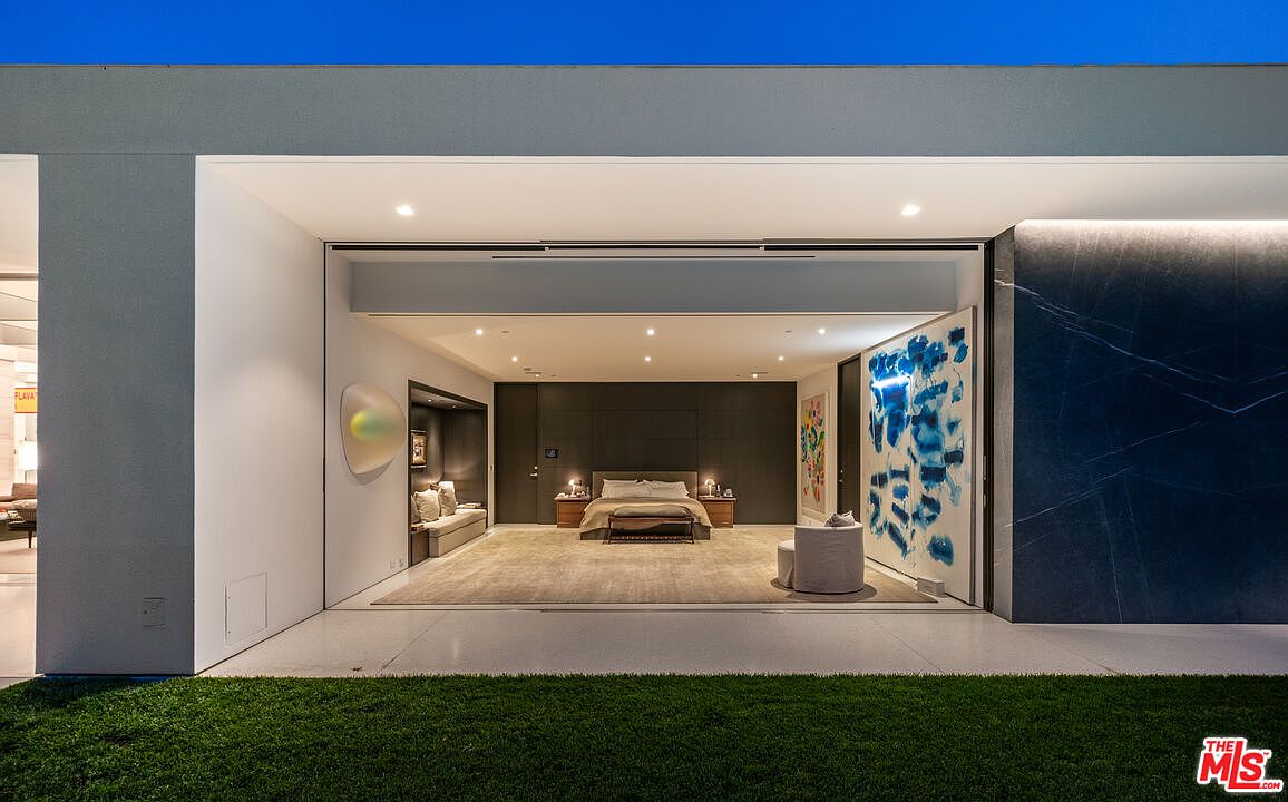This stunning primary bedroom features an expansive, open-concept design that seamlessly transitions to an outdoor lawn area through floor-to-ceiling sliding glass walls. The room is anchored by a minimalist bed, flanked by warm accent lighting, and adorned with large-scale contemporary art pieces that add a vibrant touch to the sophisticated, neutral palette. The cinematic perspective emphasizes the dramatic indoor-outdoor flow, highlighting the sleek, modern architectural lines and the luxurious, gallery-like atmosphere.