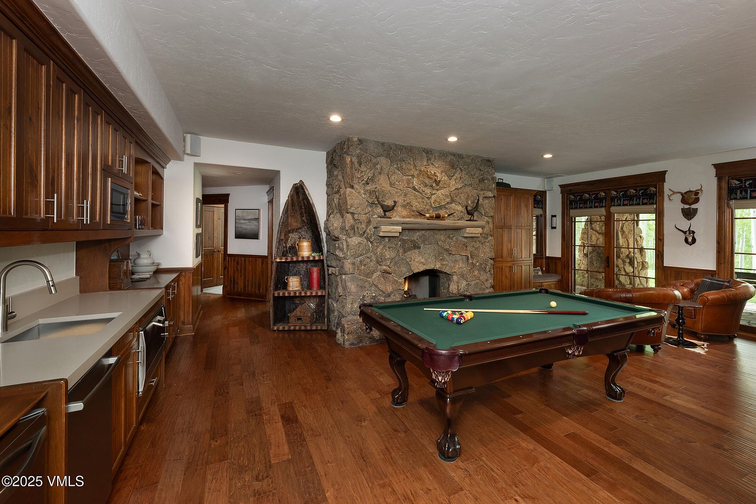 This interior shot showcases a spacious living room with a rustic yet elegant design. A stone fireplace serves as a focal point, complemented by a pool table in the foreground. The room features hardwood flooring, wood cabinetry, and comfortable leather seating, creating a warm and inviting atmosphere.