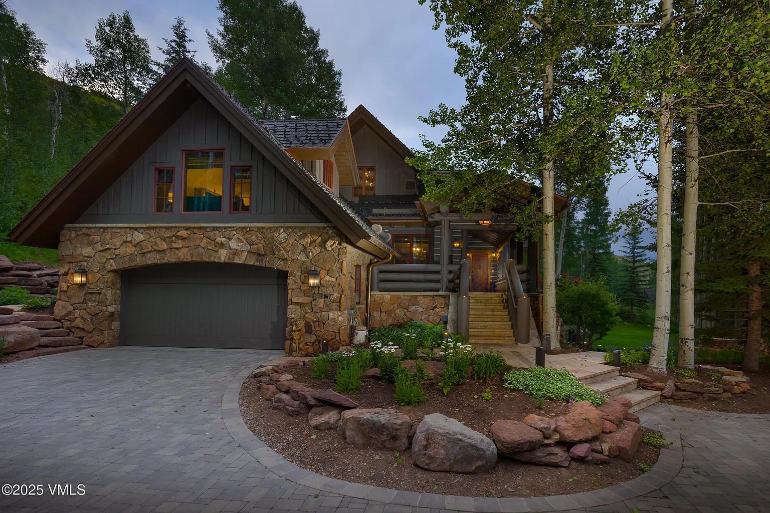 This is a front exterior view of a large, upscale home. The house features a stone-clad garage and lower level, with a contrasting gray paneled upper level. The architecture blends rustic and modern elements, and the landscaping includes a stone pathway and a well-maintained garden.