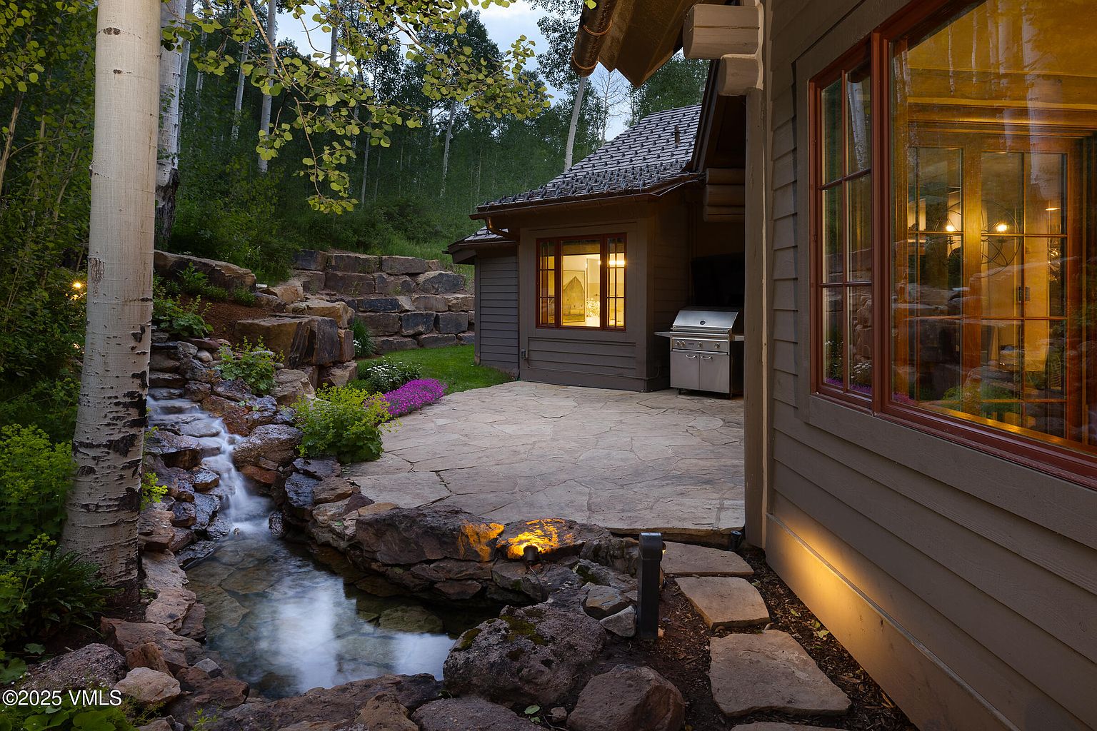 This image showcases the rear exterior of a home with a beautifully landscaped backyard. A stone patio extends from the house, featuring a built-in grill and a small waterfall feature that adds a tranquil element to the outdoor space. The home's exterior is clad in wood siding, complemented by dark-framed windows, and the surrounding landscape includes mature trees and lush greenery, creating a serene and private setting.
