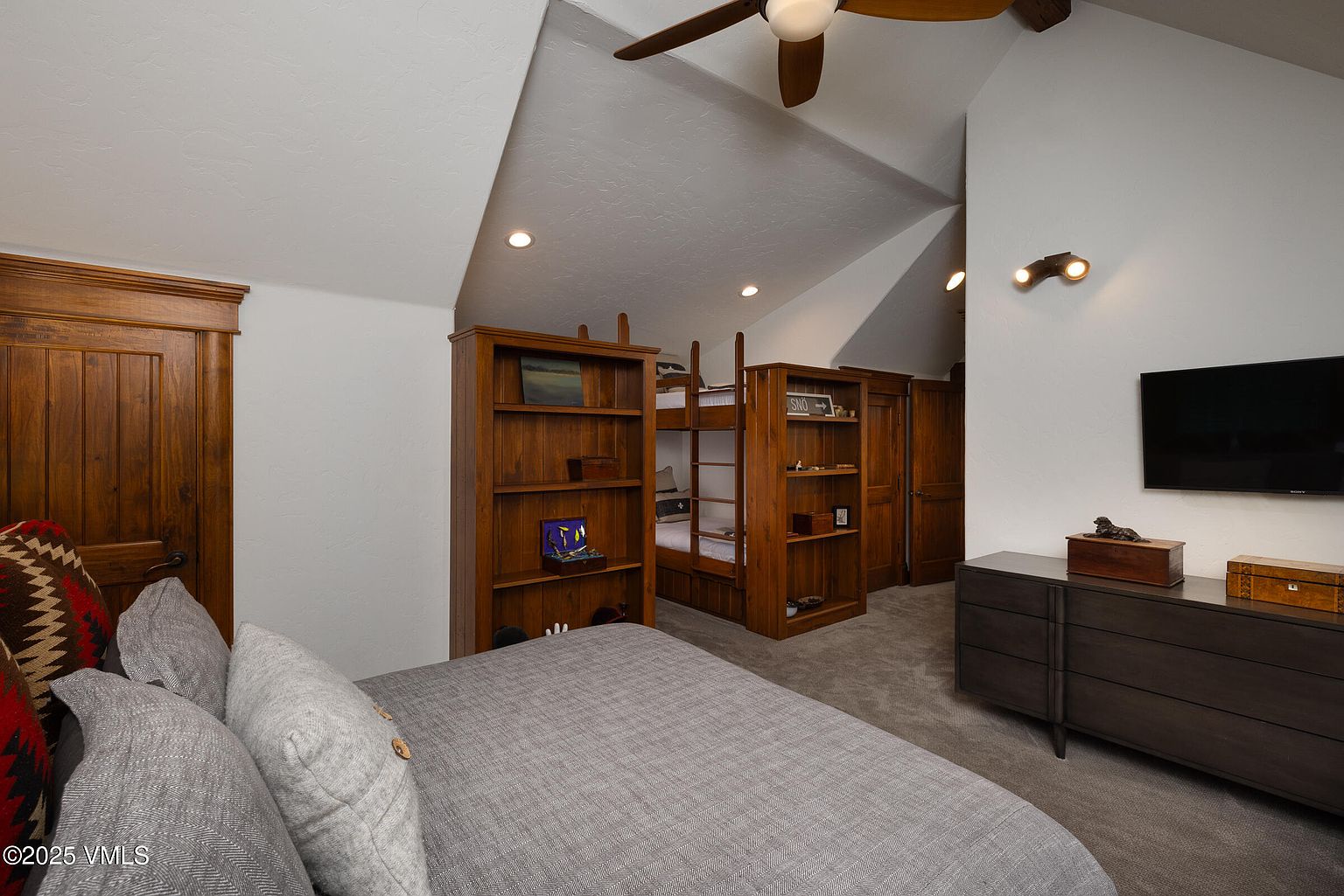 This is a cozy bedroom featuring a bunk bed built into the wall with surrounding wooden bookshelves. The room has a rustic yet modern feel, with a dark wood dresser and a flat-screen TV mounted on the wall. The bed in the foreground is neatly made with gray bedding and decorative pillows, creating an inviting atmosphere.