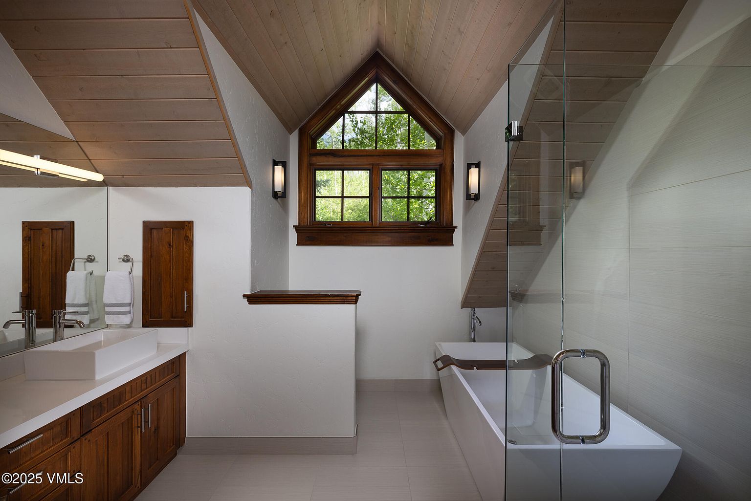 This is a well-lit primary bathroom featuring a modern aesthetic with a rustic touch. The bathroom includes a vanity with a rectangular sink, wooden cabinets, and a large mirror. A freestanding bathtub is enclosed by a glass shower screen, and a triangular window provides natural light, complemented by sconces on either side.