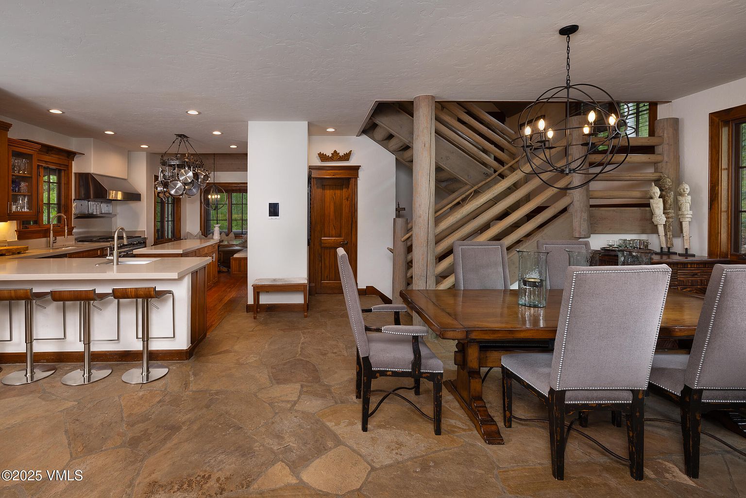 This interior shot showcases a dining area adjacent to a kitchen, featuring a rustic yet elegant design. The dining table is surrounded by upholstered chairs, and a unique orb chandelier hangs above. A wooden staircase and stone flooring add to the room's character, creating a warm and inviting atmosphere.