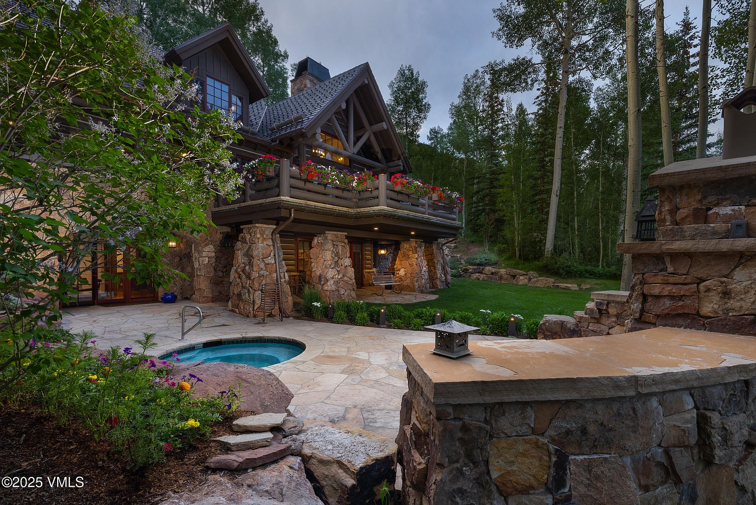 This image showcases a luxurious outdoor living space with a stone patio, a built-in hot tub, and a balcony adorned with vibrant flower boxes. The architecture features a blend of wood and stone, creating a rustic yet elegant aesthetic. The surrounding landscape includes lush greenery and mature trees, enhancing the property's privacy and appeal.