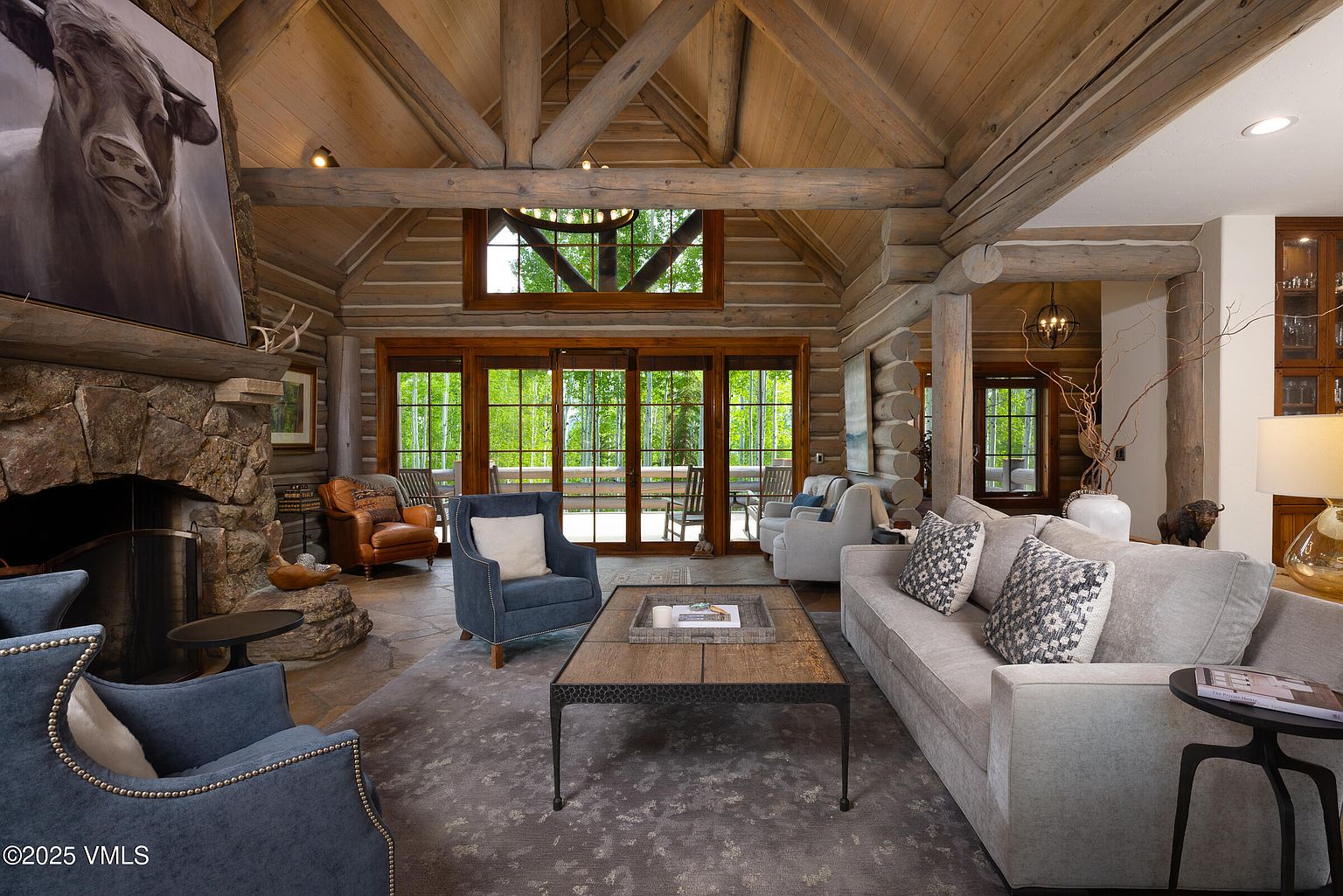 This is an interior shot of a rustic living room featuring a stone fireplace with a large painting above it, exposed wooden beams, and large windows overlooking a lush green forest. The room is furnished with comfortable seating, including a gray sofa and blue armchairs, arranged around a dark metal and wood coffee table on a patterned rug. The overall impression is one of warmth, luxury, and connection to nature.