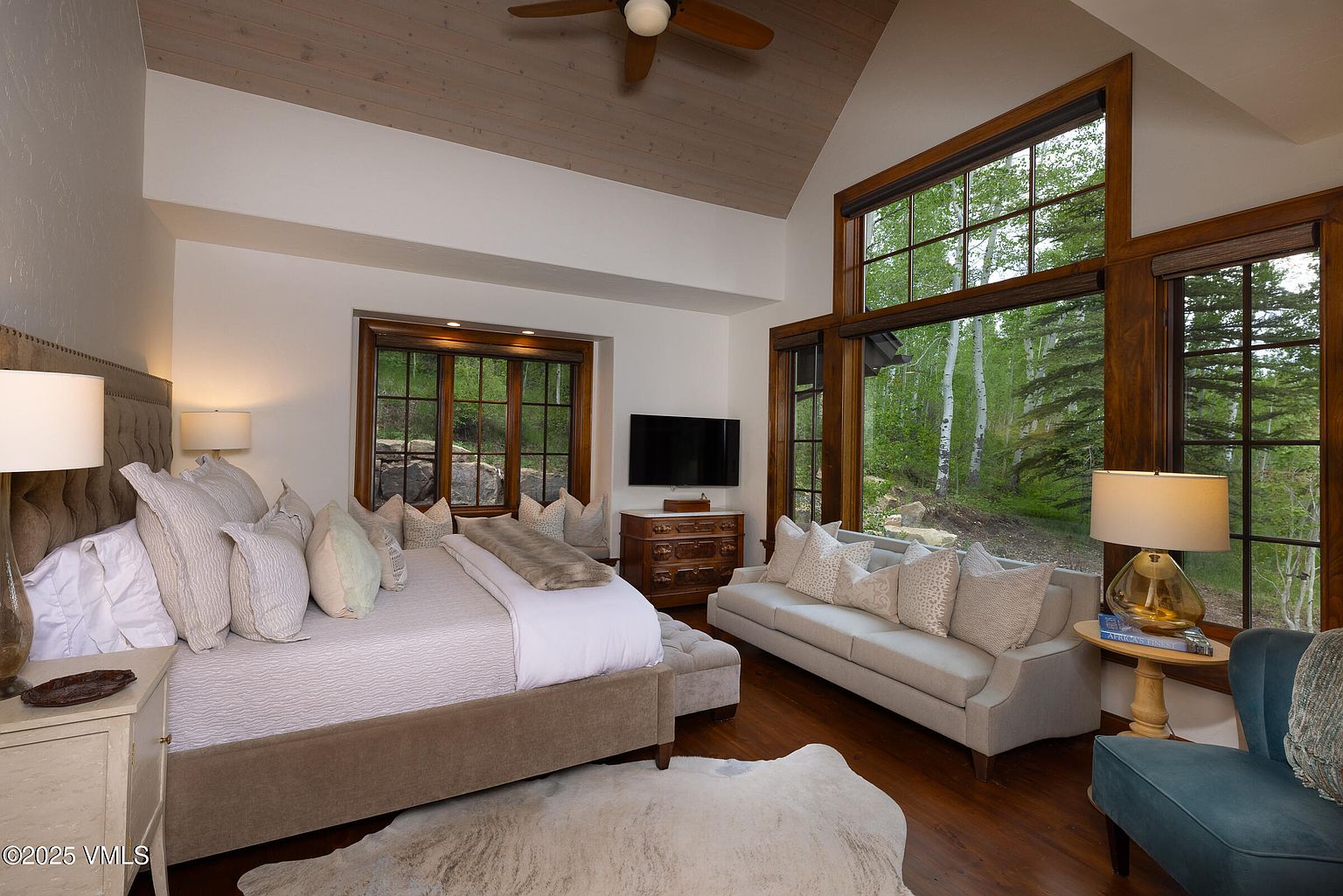 This is a luxurious primary bedroom featuring a king-size bed with a tufted headboard, multiple decorative pillows, and a cozy throw. Large windows with dark wood frames offer views of the surrounding trees, and a comfortable sofa provides a seating area. The room is well-lit with a combination of natural light and lamps, creating a warm and inviting atmosphere.