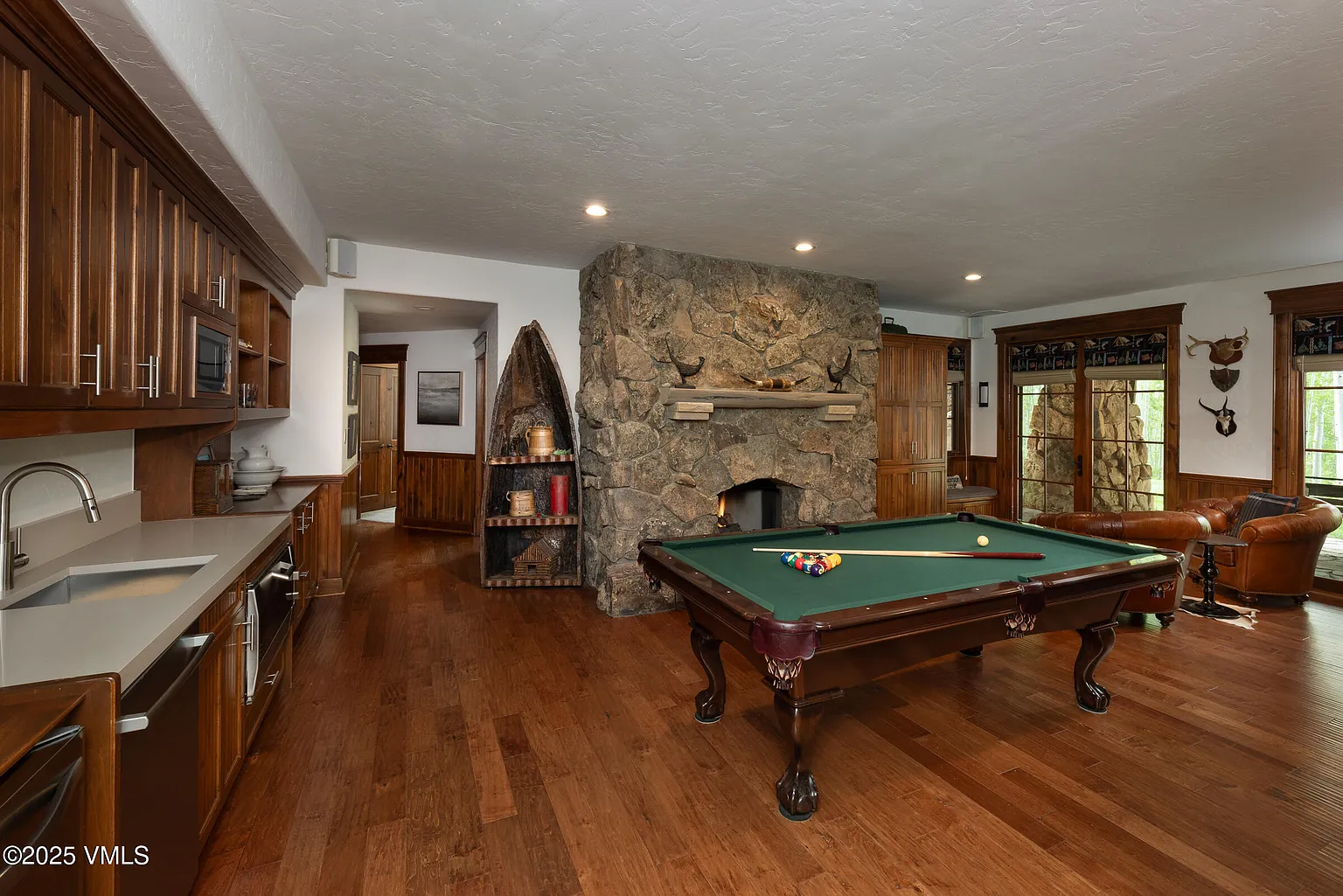 This is an interior shot of a living room featuring a pool table, a stone fireplace, and wooden accents. The room has hardwood floors and built-in wooden cabinetry, indicative of a rustic and luxurious design. The overall impression is that of a comfortable and well-appointed recreational space.