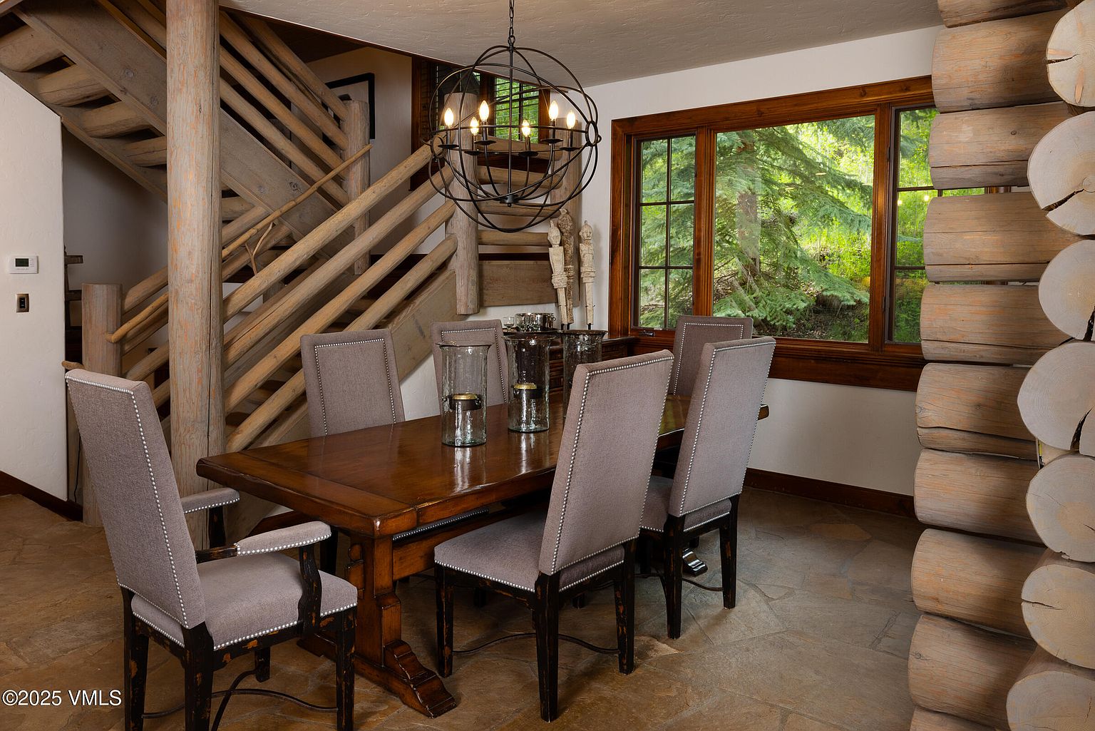 This is an interior shot of a dining room featuring a wooden dining table with six upholstered chairs. A unique spherical chandelier hangs above the table, and a window provides natural light and a view of the outdoors. The room also showcases a rustic wooden staircase and log wall, adding to the cabin-like ambiance.
