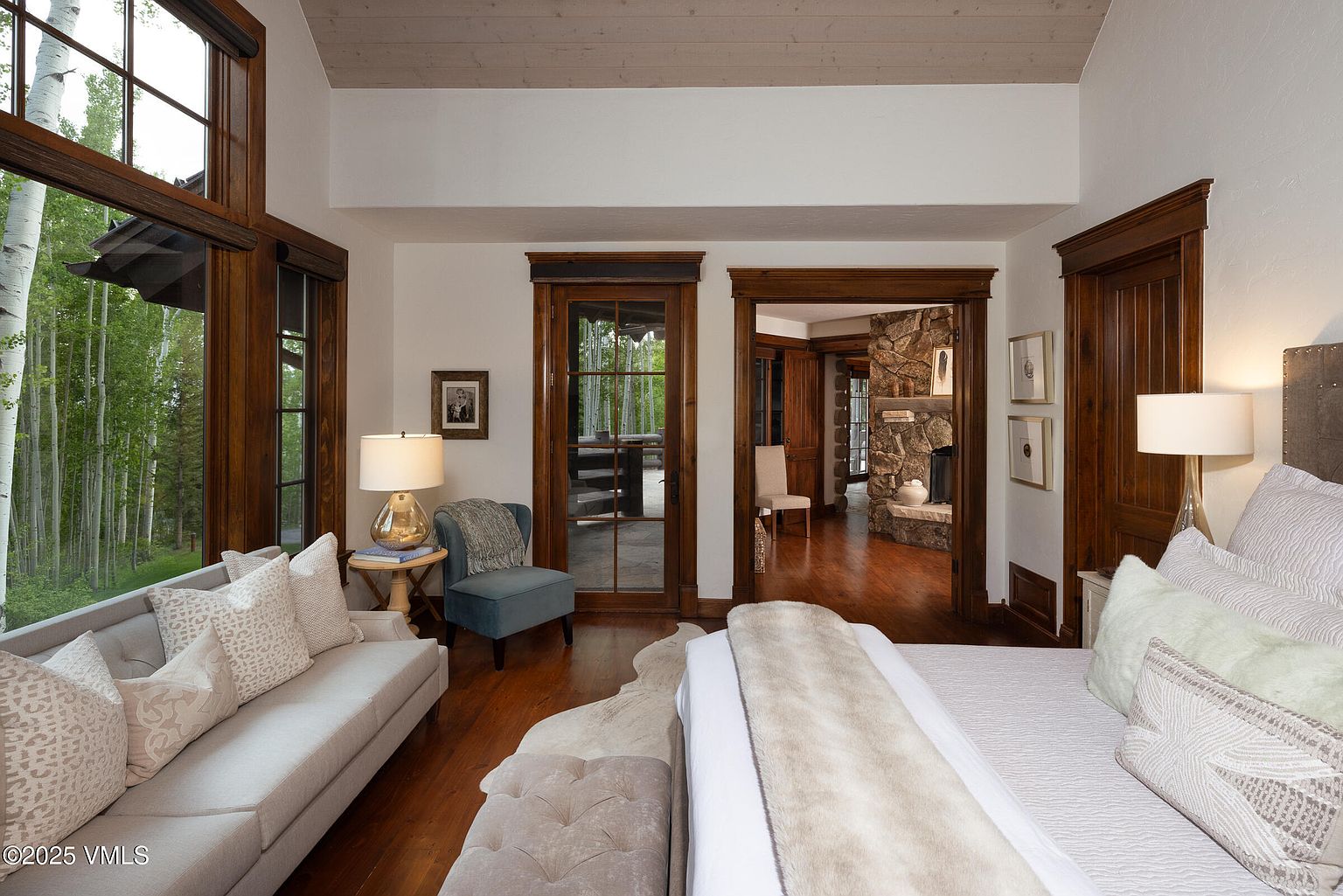 This is a primary bedroom featuring a sitting area with a light gray sofa and a blue accent chair. The room has hardwood floors, wood-trimmed windows offering a view of the forest, and a doorway leading to another room with a stone fireplace. The bed is neatly made with neutral-toned bedding, creating a serene and inviting atmosphere.