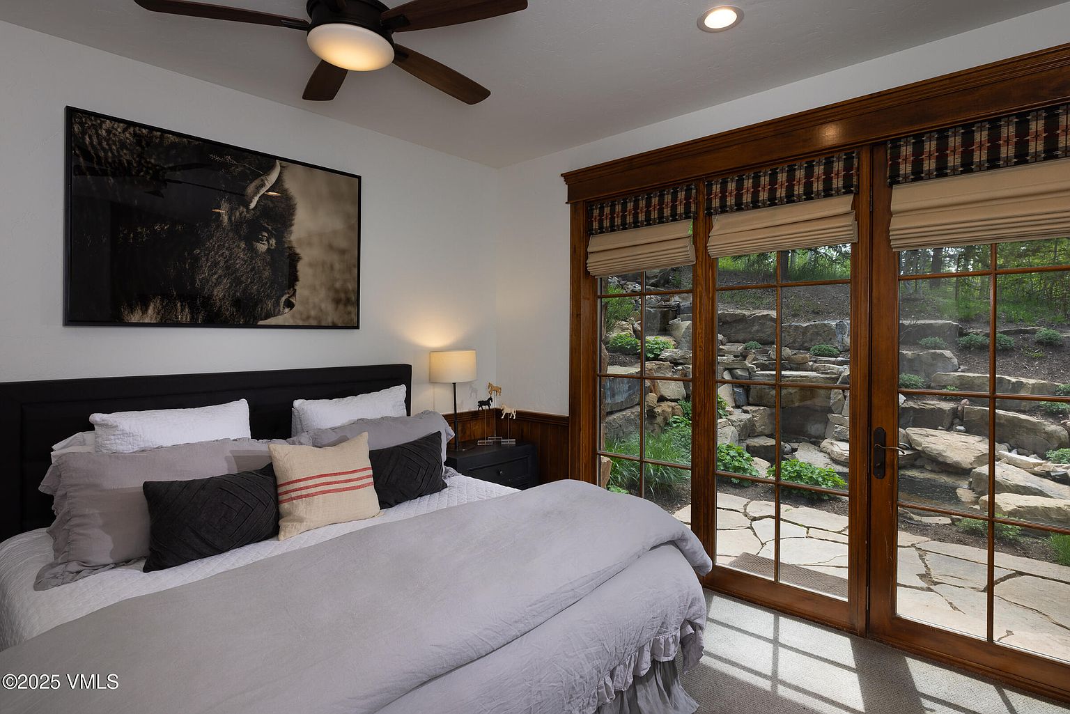 This is a cozy bedroom featuring a comfortable bed with multiple pillows and a gray comforter. A large framed picture of a bison hangs above the bed, adding a rustic touch. The room has a door leading to an outdoor area with a rock garden, providing a serene view and natural light.