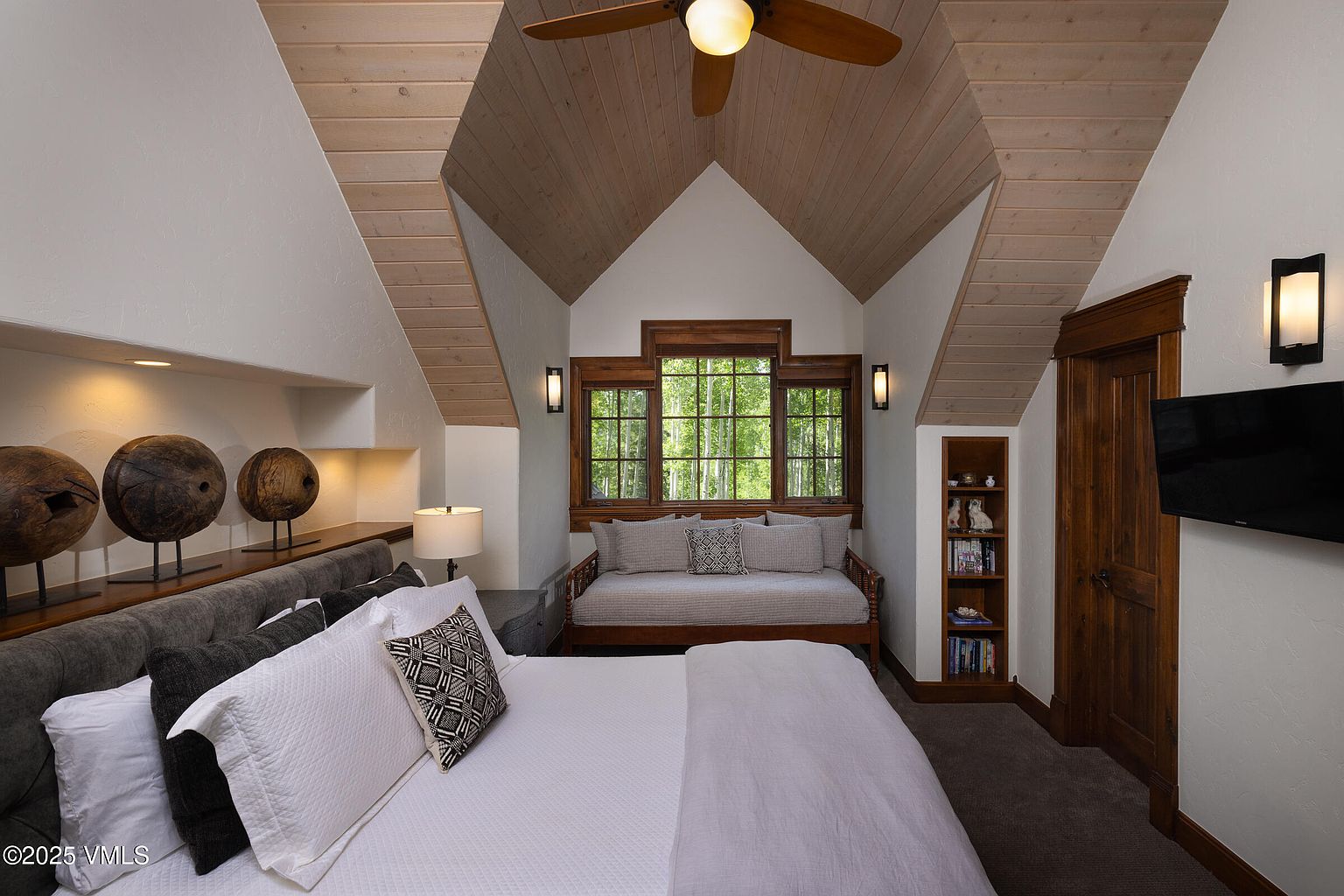 This is a cozy bedroom featuring a unique architectural design with a sloped ceiling and wooden accents. The room includes a bed with white linens and decorative pillows, a window seat with a view of trees, and built-in shelving. The overall impression is warm and inviting, perfect for a guest room or secondary bedroom.