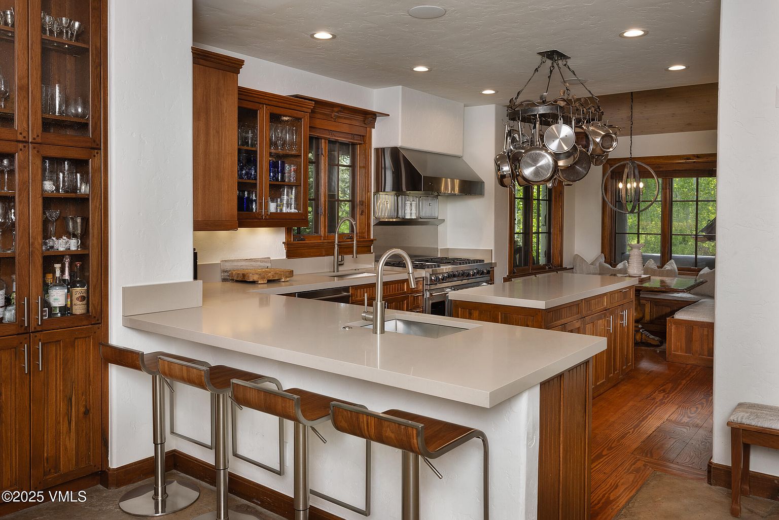 This is a well-appointed kitchen featuring custom wood cabinetry with glass-fronted upper cabinets, light countertops, and stainless steel appliances. A central island with bar seating provides a gathering space, while a pot rack hangs above the cooking area. The hardwood flooring and natural light create a warm and inviting atmosphere.