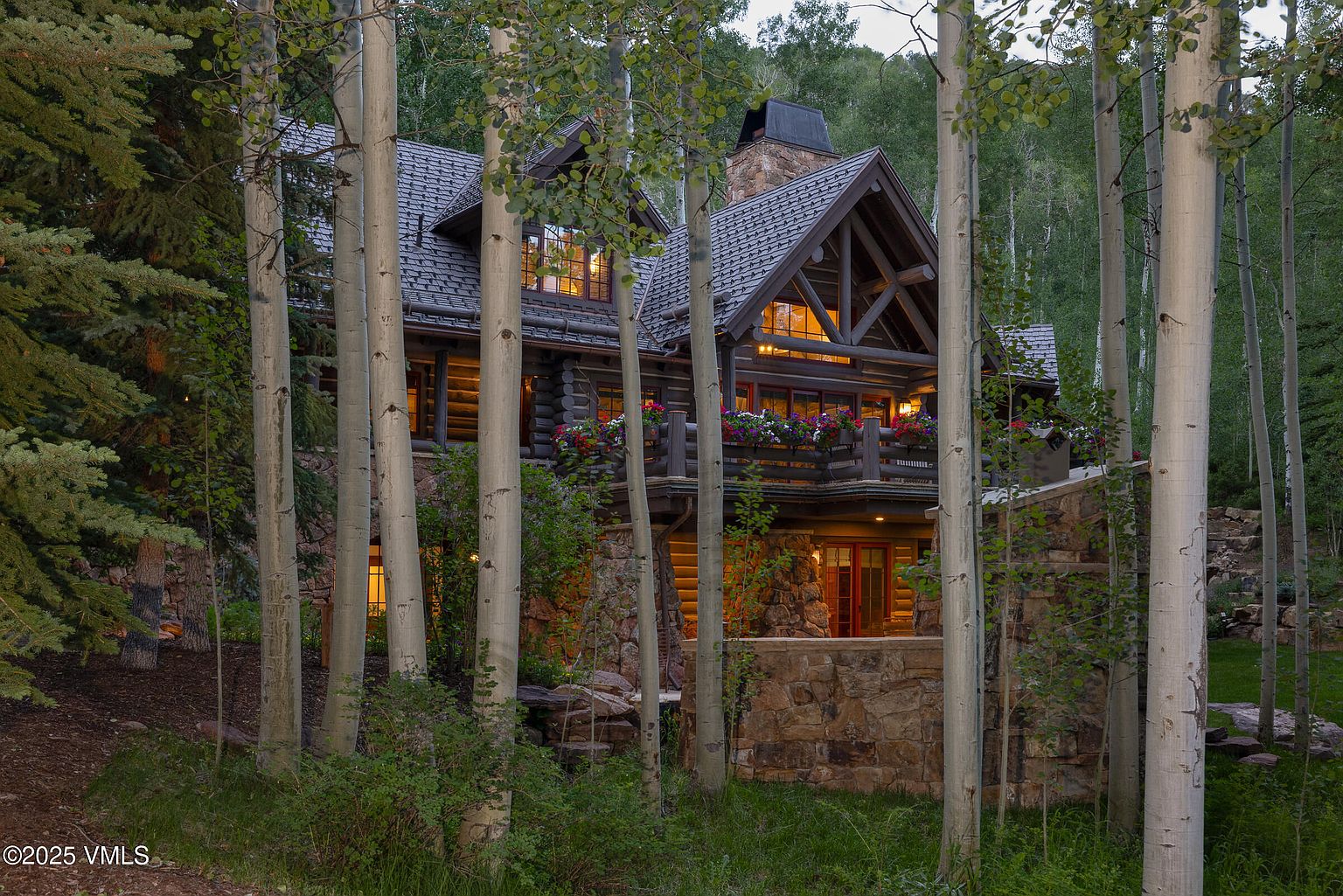 This image showcases the front exterior of a luxurious log cabin home nestled among aspen trees. The home features a stone foundation, a balcony with flower boxes, and a steeply pitched roof with a stone chimney. Warm light emanates from the windows, creating an inviting and cozy atmosphere.